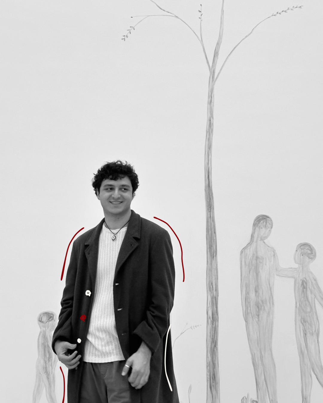 A smiling young man with curly hair wearing a coat and white sweater standing in front of a wall with a large drawing of a tree and two figures.