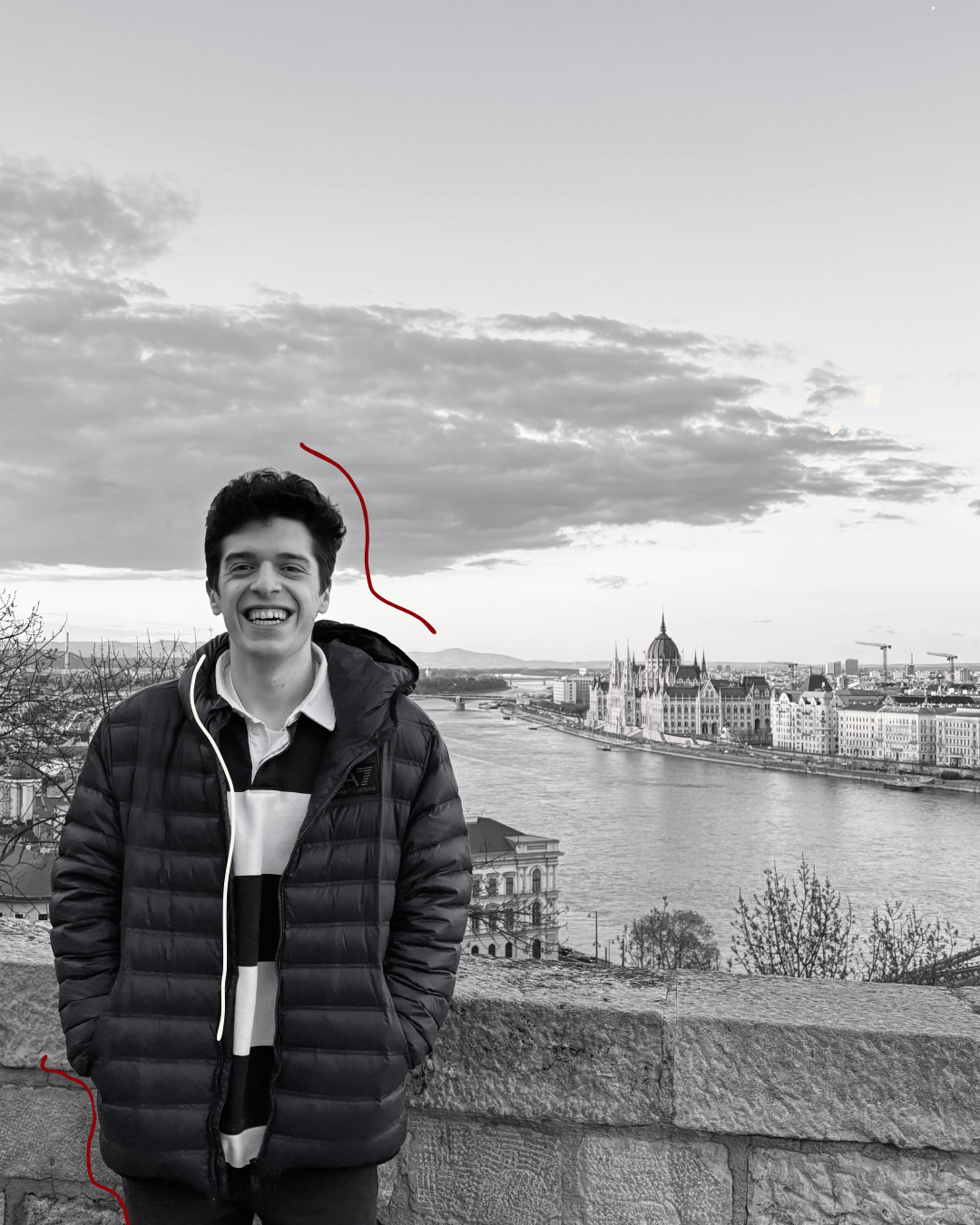 Young man smiling outdoors near a stone railing, with a cityscape and river in the background, black and white photograph.