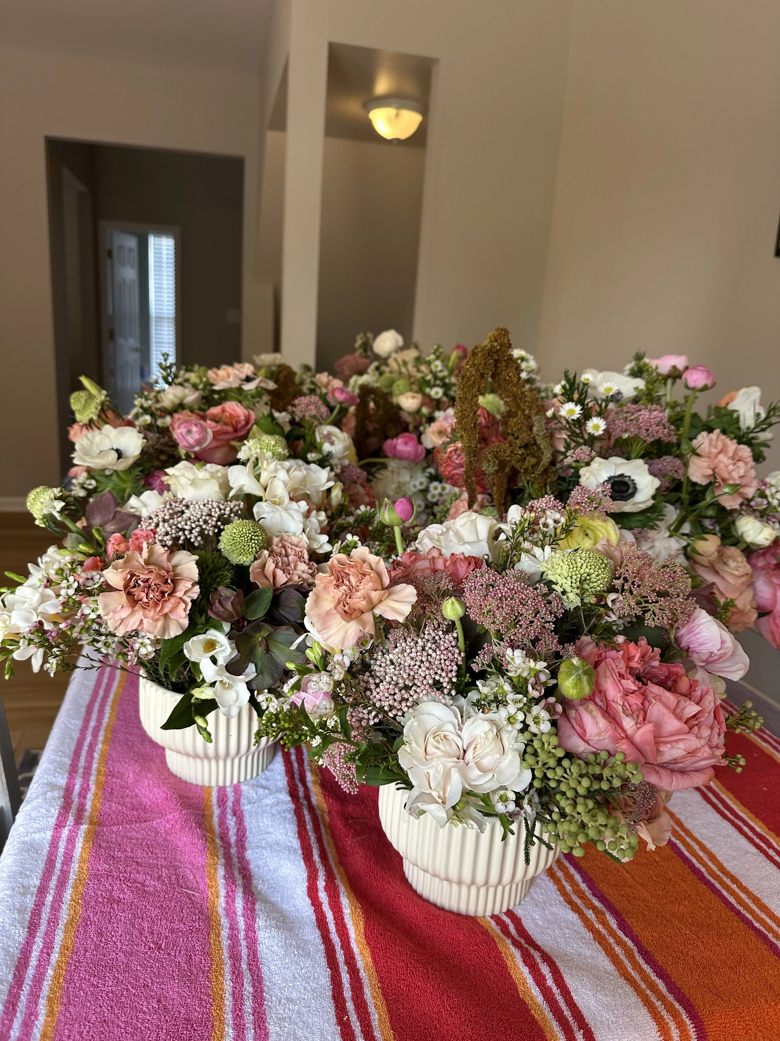 Two floral bouquets with pink, white, and blush flowers in white vases on a striped pink and red tablecloth in a room with beige walls.