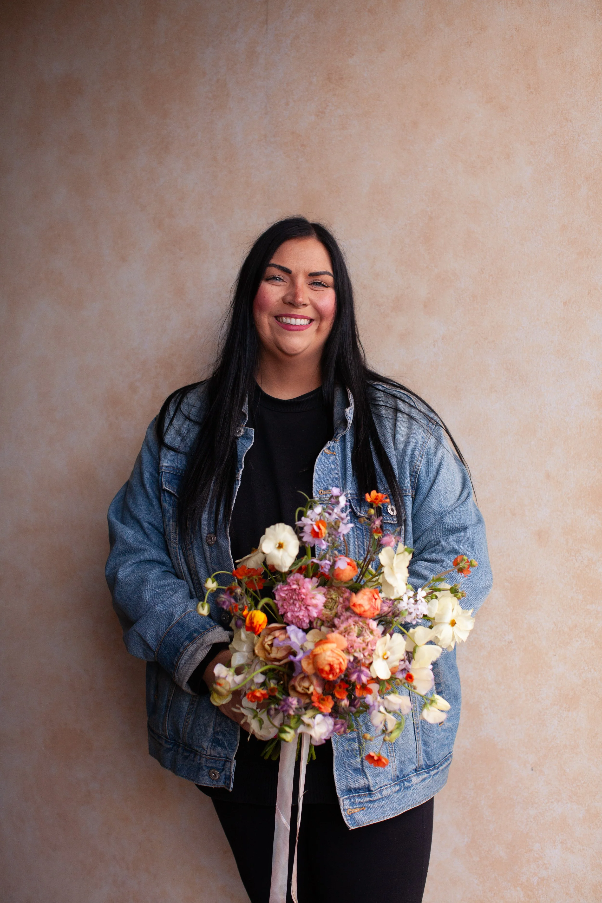 A woman with long black hair smiling and holding a colorful bouquet of flowers, standing against a beige textured wall.