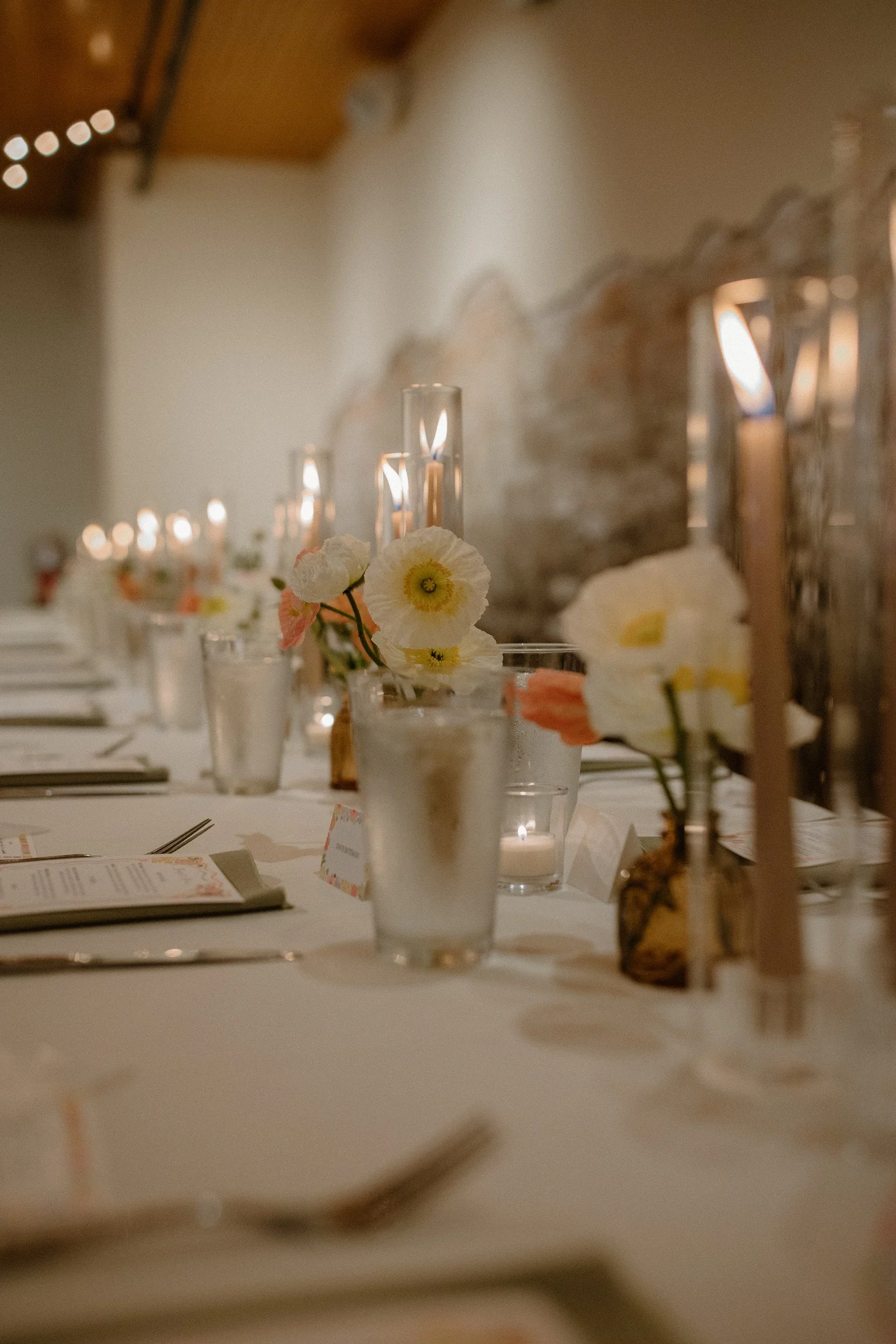 A close-up of a decorated table with flowers in small vases, candles, and place cards, set for a formal event or celebration in a rustic venue with wooden beams and a white wall.