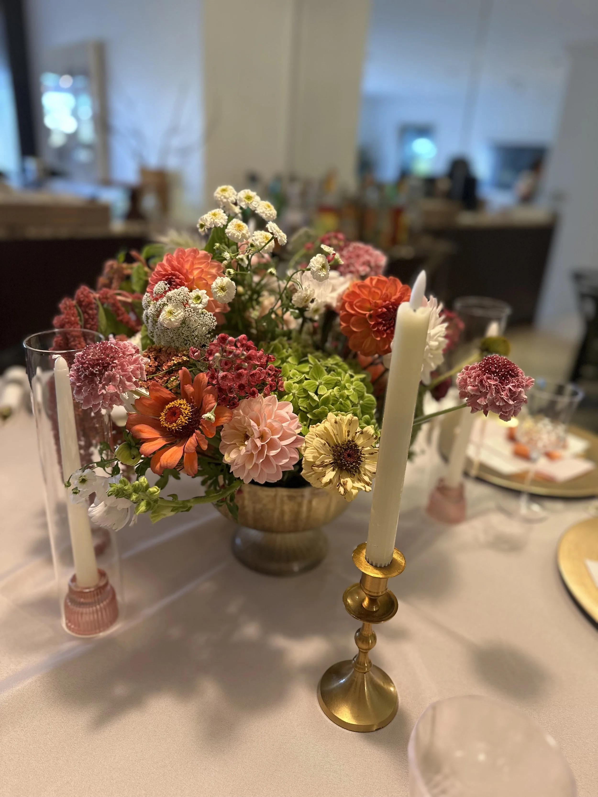 A floral centerpiece with pink, red, orange, and white flowers on a dining table, accompanied by white candles in gold candlesticks.