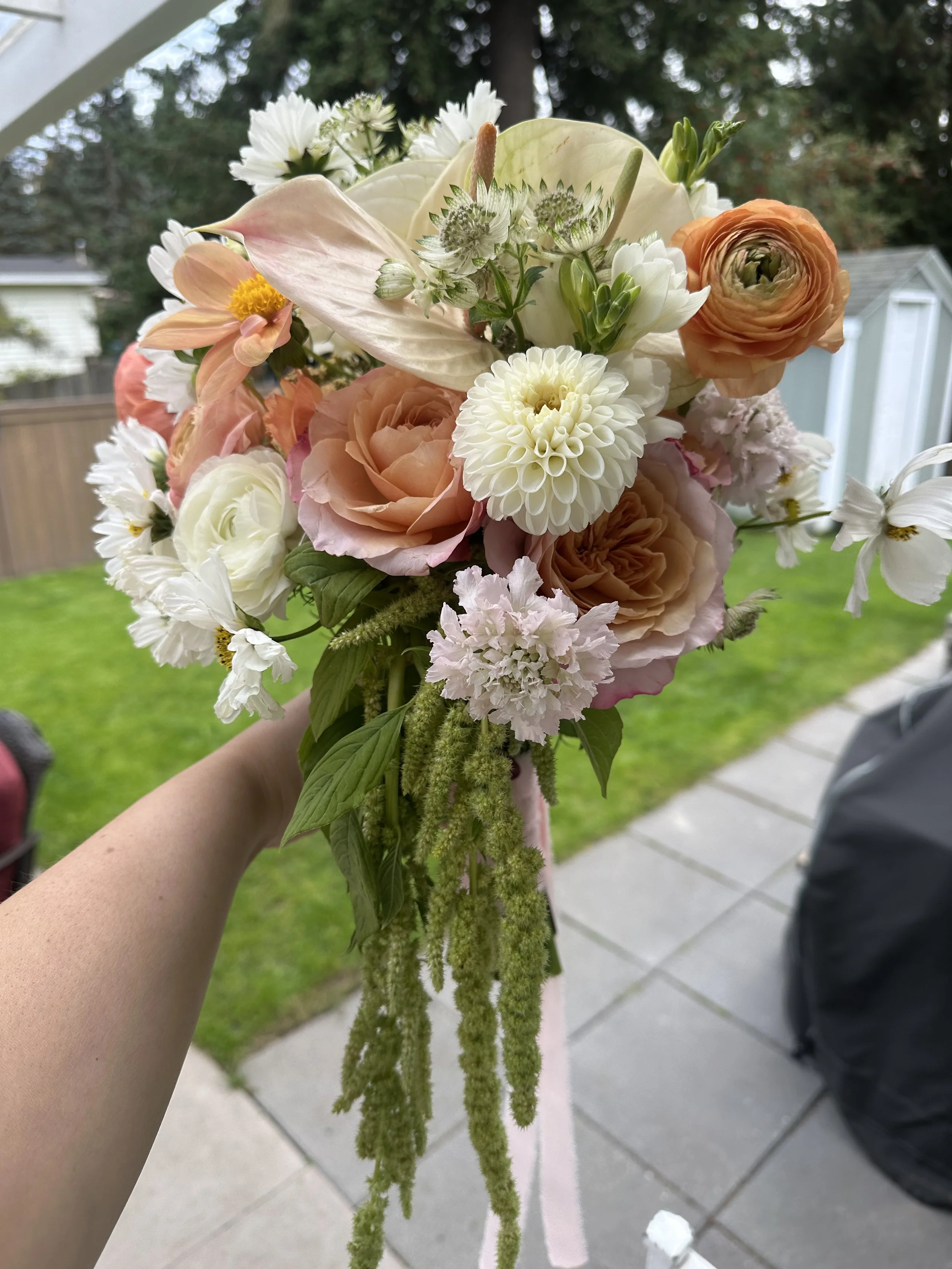 A bouquet of various flowers including roses, dahlias, and anthurium, with greenery, held by a person's hand outside near a garden.