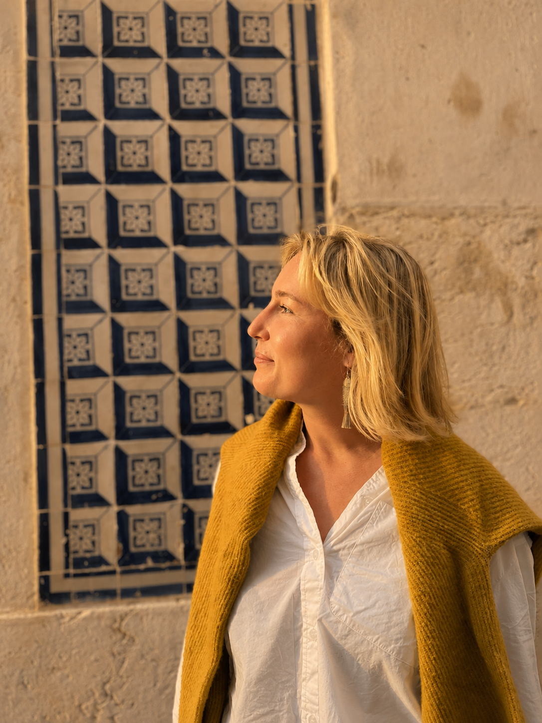 A woman with blonde hair, wearing a white shirt and a yellow jacket, standing against a tiled wall with a geometric pattern, smiling and looking to the left.