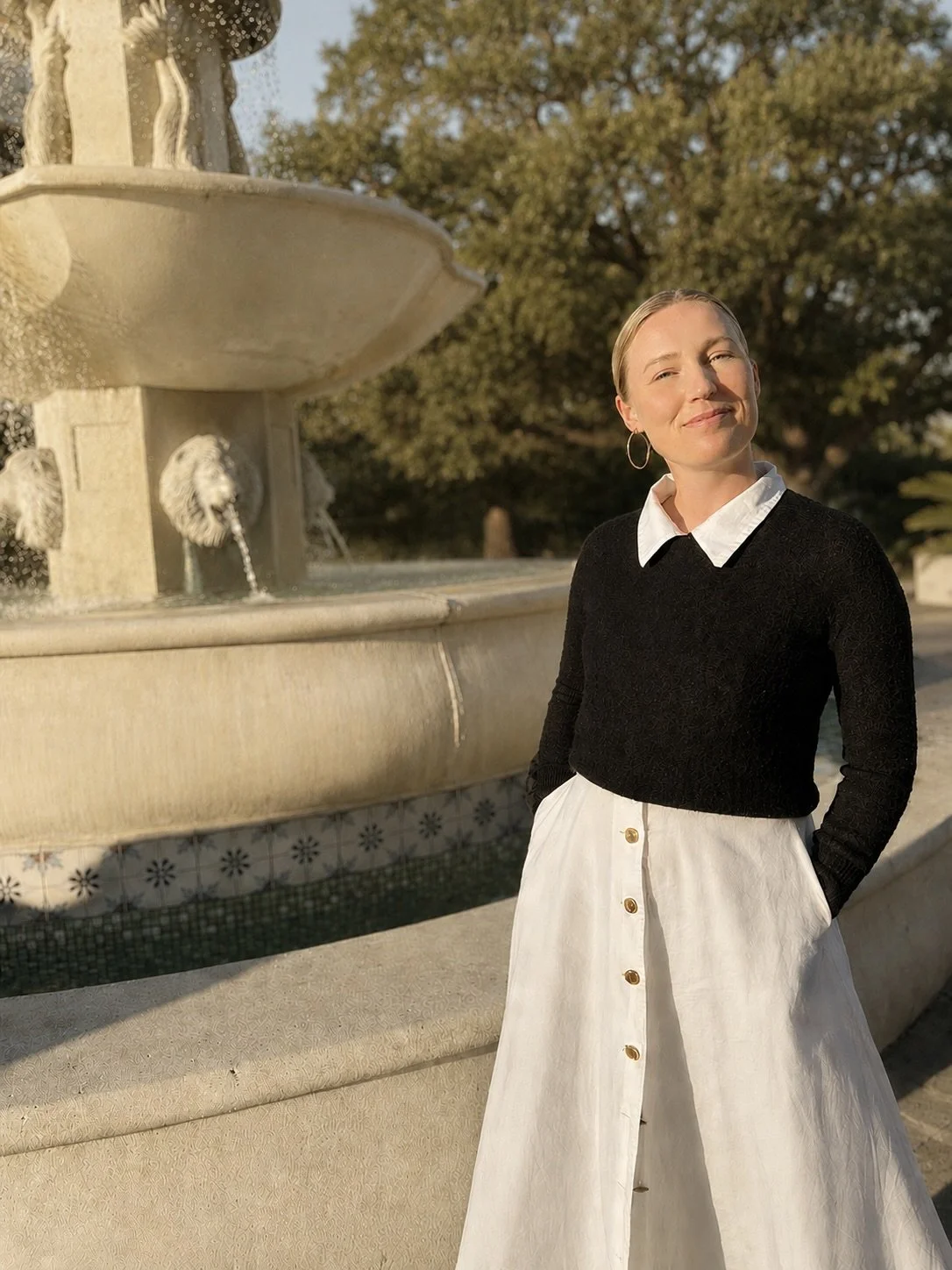 A woman in a black sweater with a white collared shirt underneath and a white skirt with buttons stands beside a stone fountain with lion heads in an outdoor setting with trees in the background.