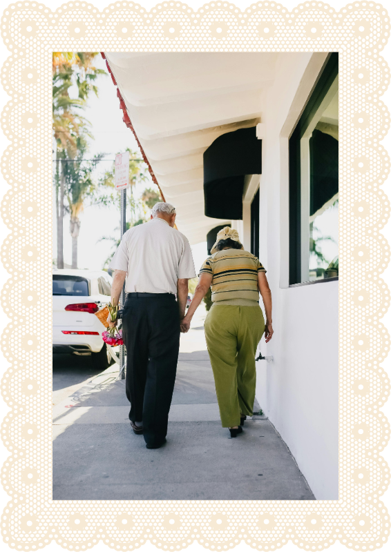 An elderly couple walking hand in hand on a sidewalk in a sunny area.