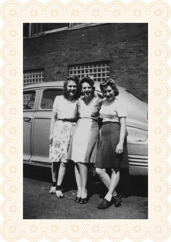 Black and white photo of three women standing together in front of a vintage car, smiling, with a brick building in the background.
