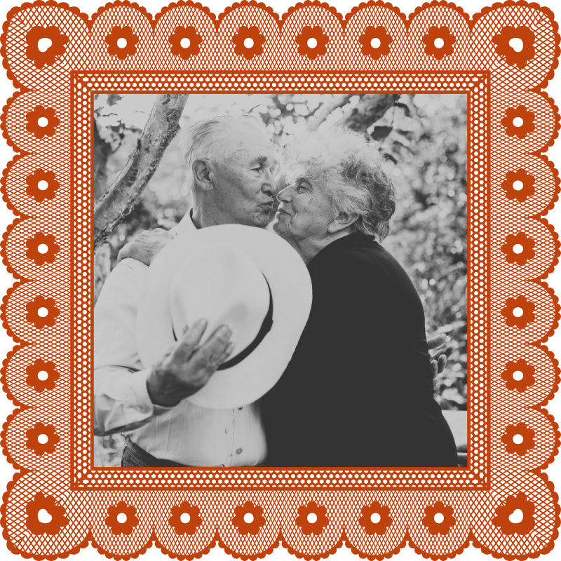 An elderly man and woman embracing outdoors, with the man holding a white hat and face-to-face at close distance.