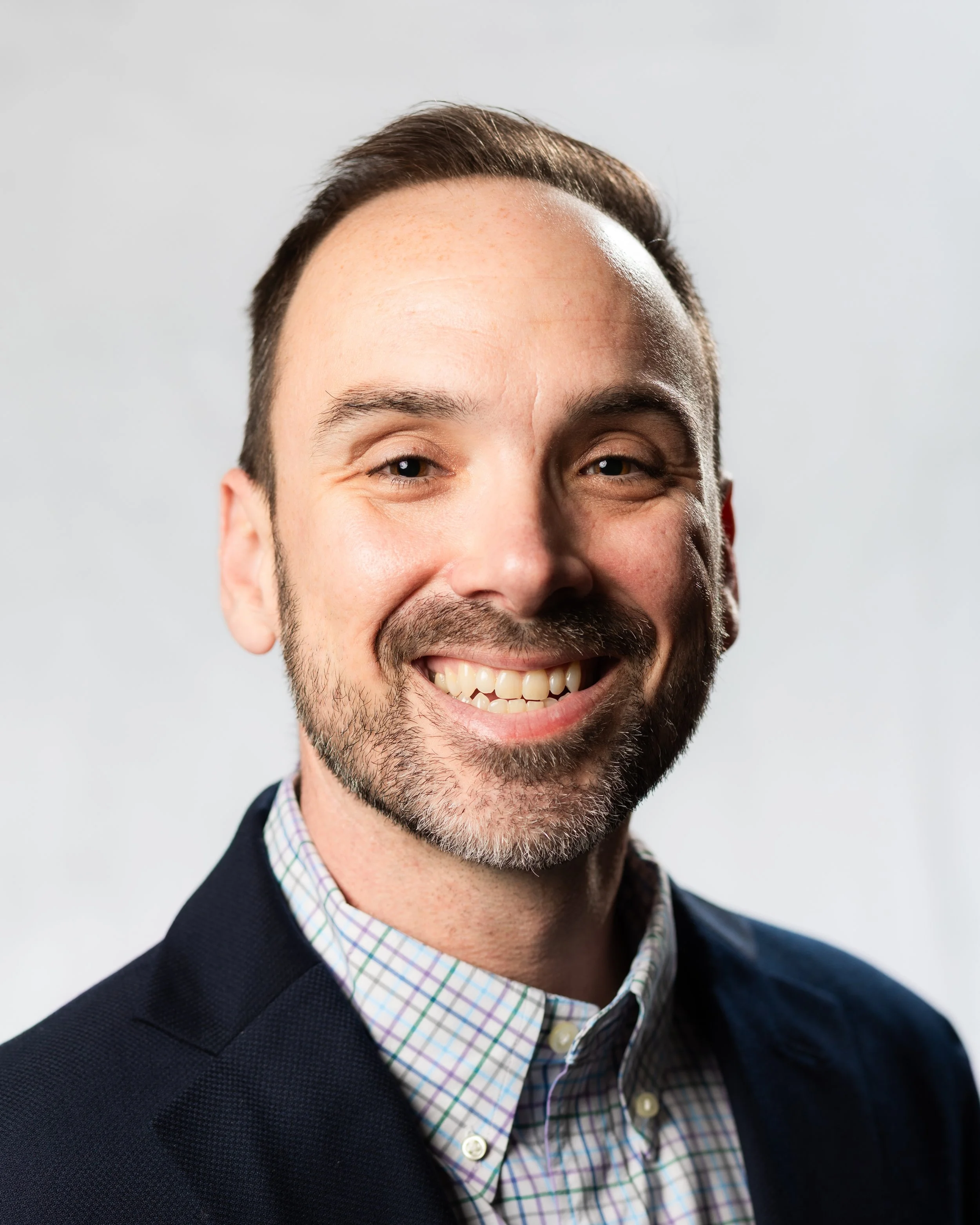 Portrait of a smiling man with short brown hair, light beard, wearing a dark blazer and checkered shirt, against a plain light background.