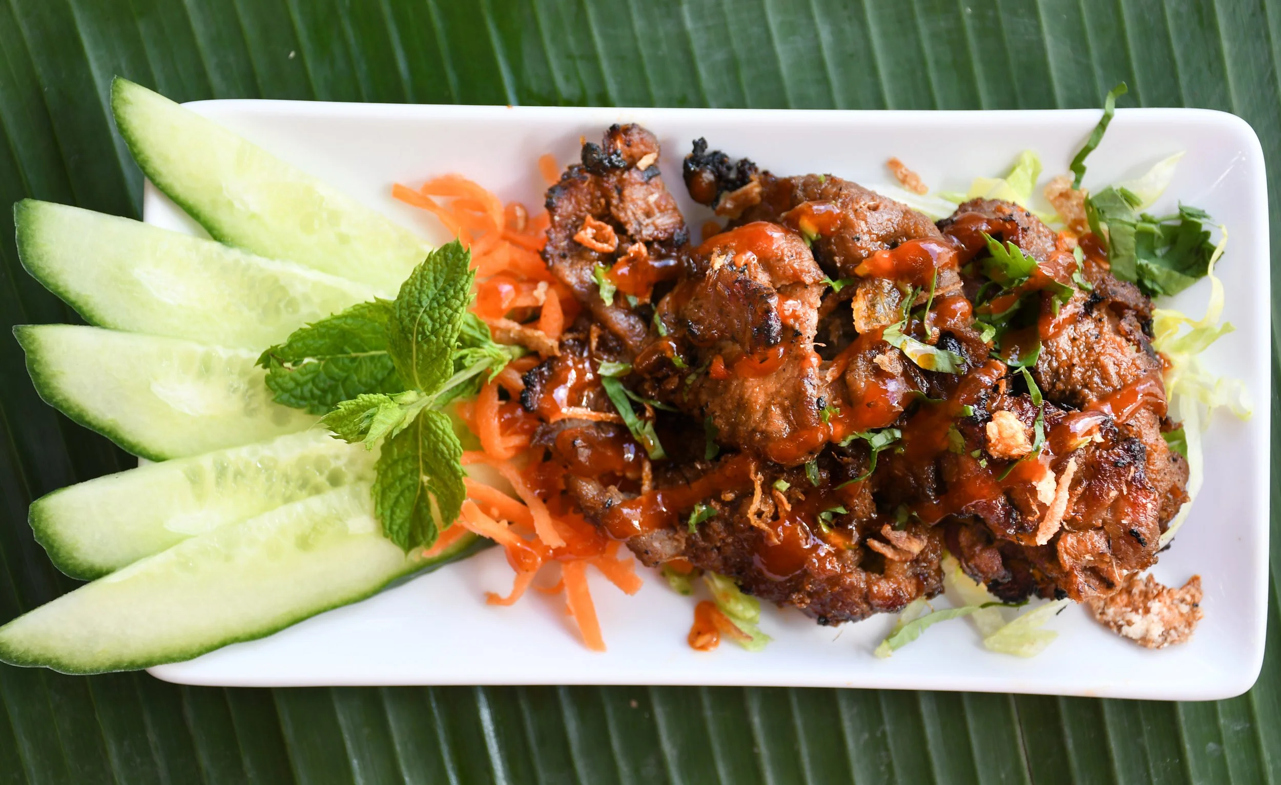 Fried meat pieces topped with sauce and herbs, served with cucumber slices, shredded carrots, and lettuce on a white rectangular plate, garnished with mint leaves.