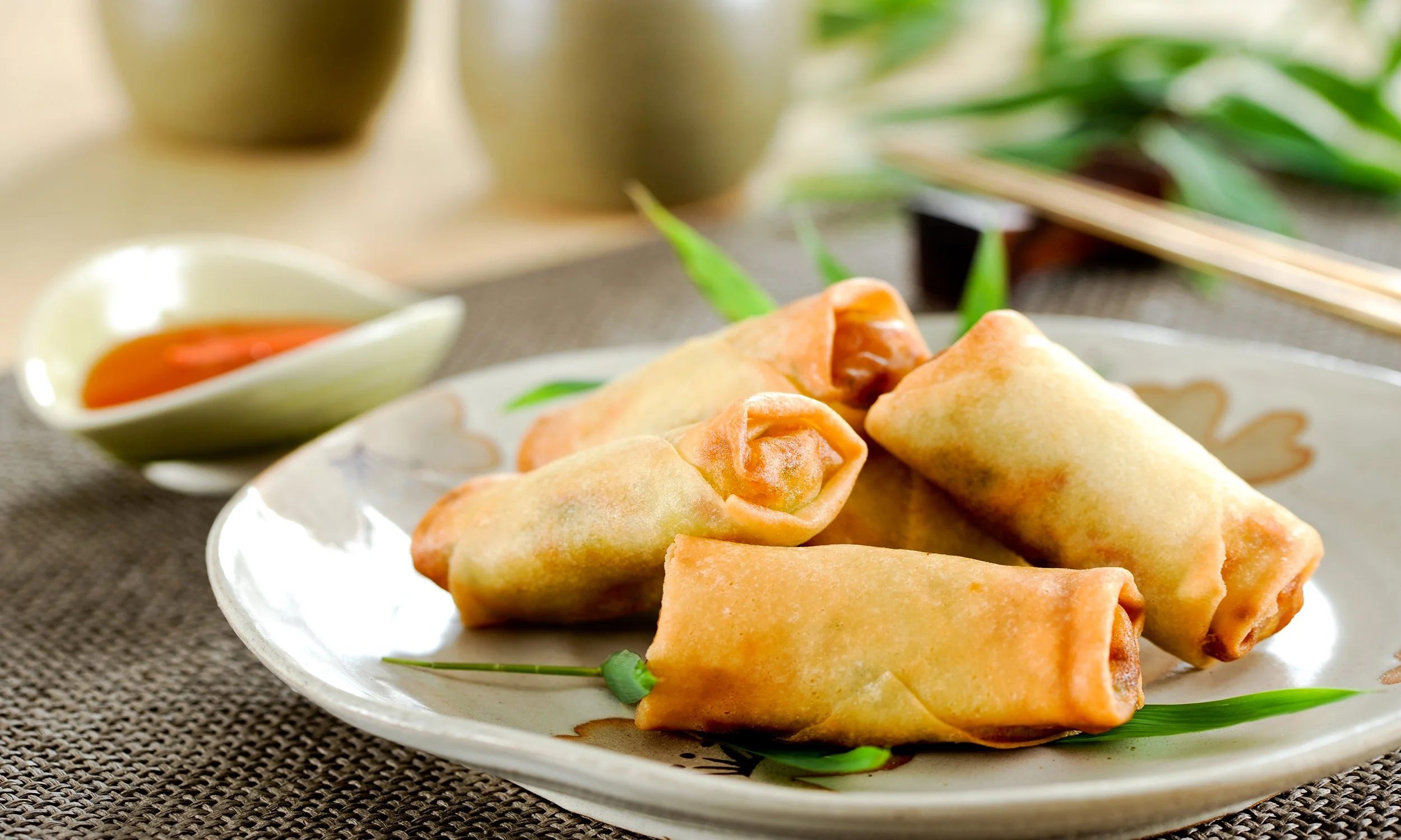 Six spring rolls on a decorative plate with dipping sauce in a bowl, bamboo leaves in the background, and chopsticks visible at the top right.