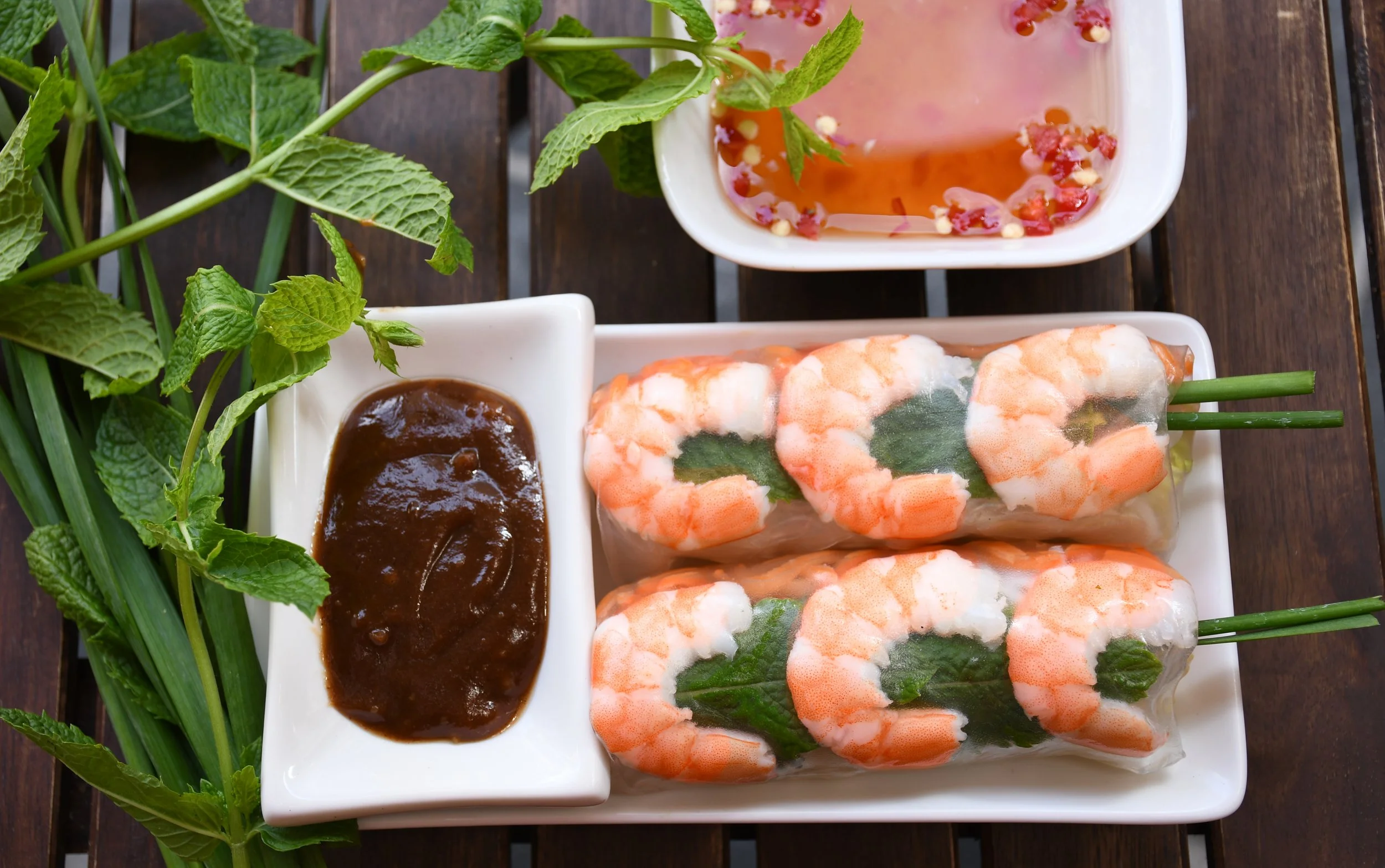 Fresh spring rolls filled with shrimp, herbs, and rice noodles with a side of dipping sauces, garnished with mint leaves and green onions.