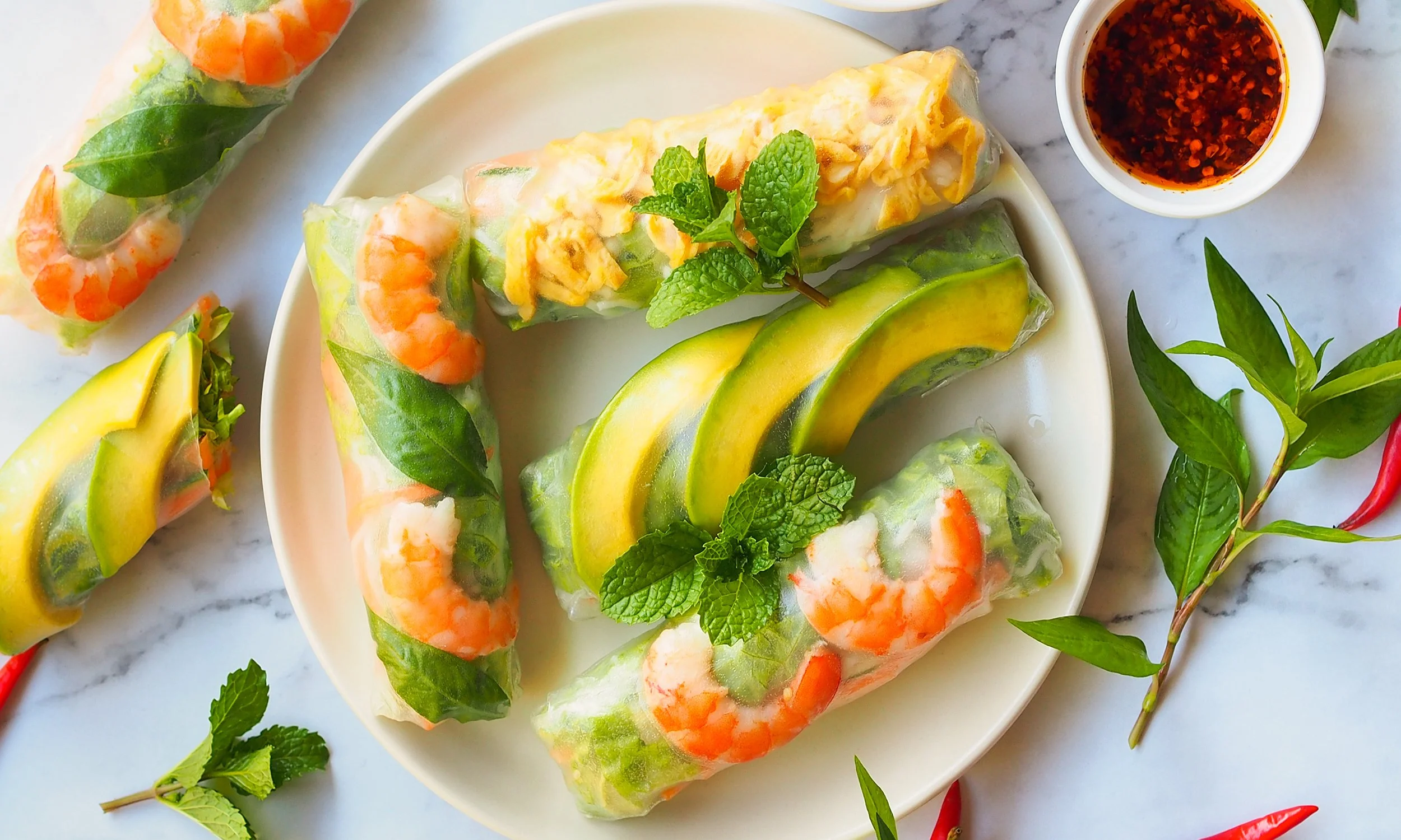 Vietnamese spring rolls with shrimp, fresh herbs, and sliced avocado on a white plate, served with a small bowl of dipping sauce, garnished with mint and basil leaves.