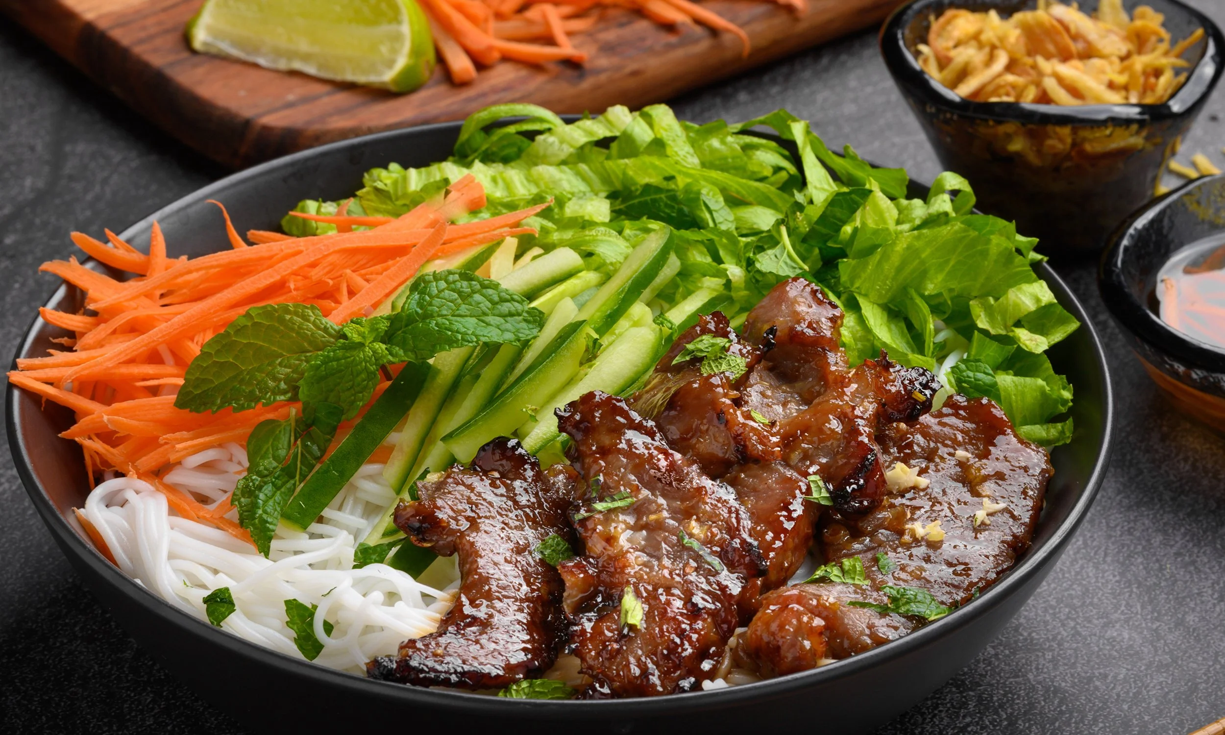 Bowl of Asian noodle salad with shredded carrots, cucumber, lettuce, grilled meat, and fresh mint leaves on top.