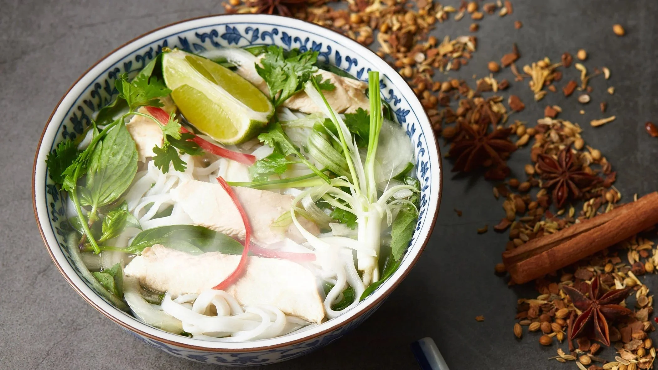 A bowl of Vietnamese pho with sliced chicken, rice noodles, herbs, and lime wedges on a dark surface with star anise and spices scattered nearby.