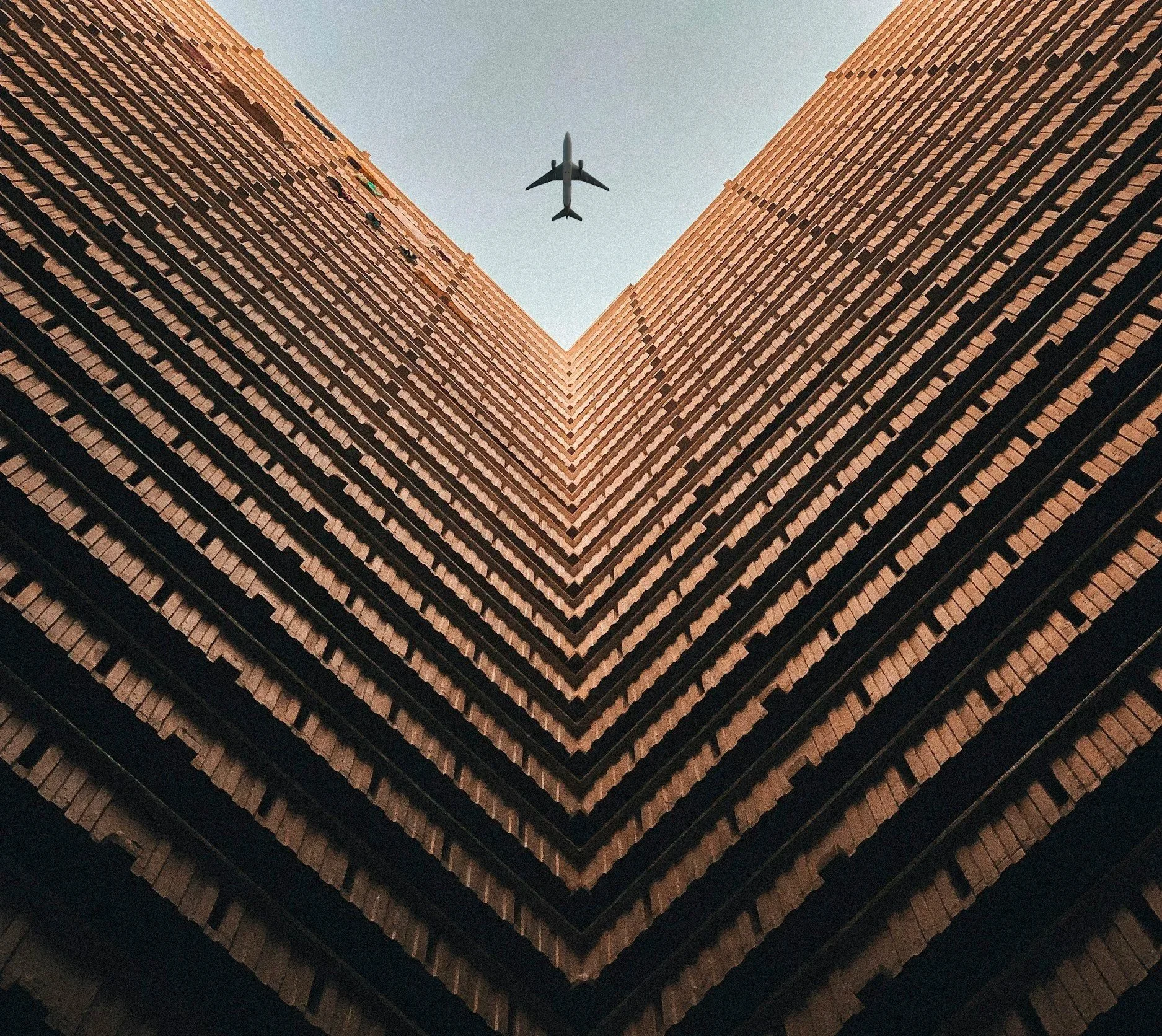 Looking up at the corner of tall brick buildings with an airplane flying overhead.