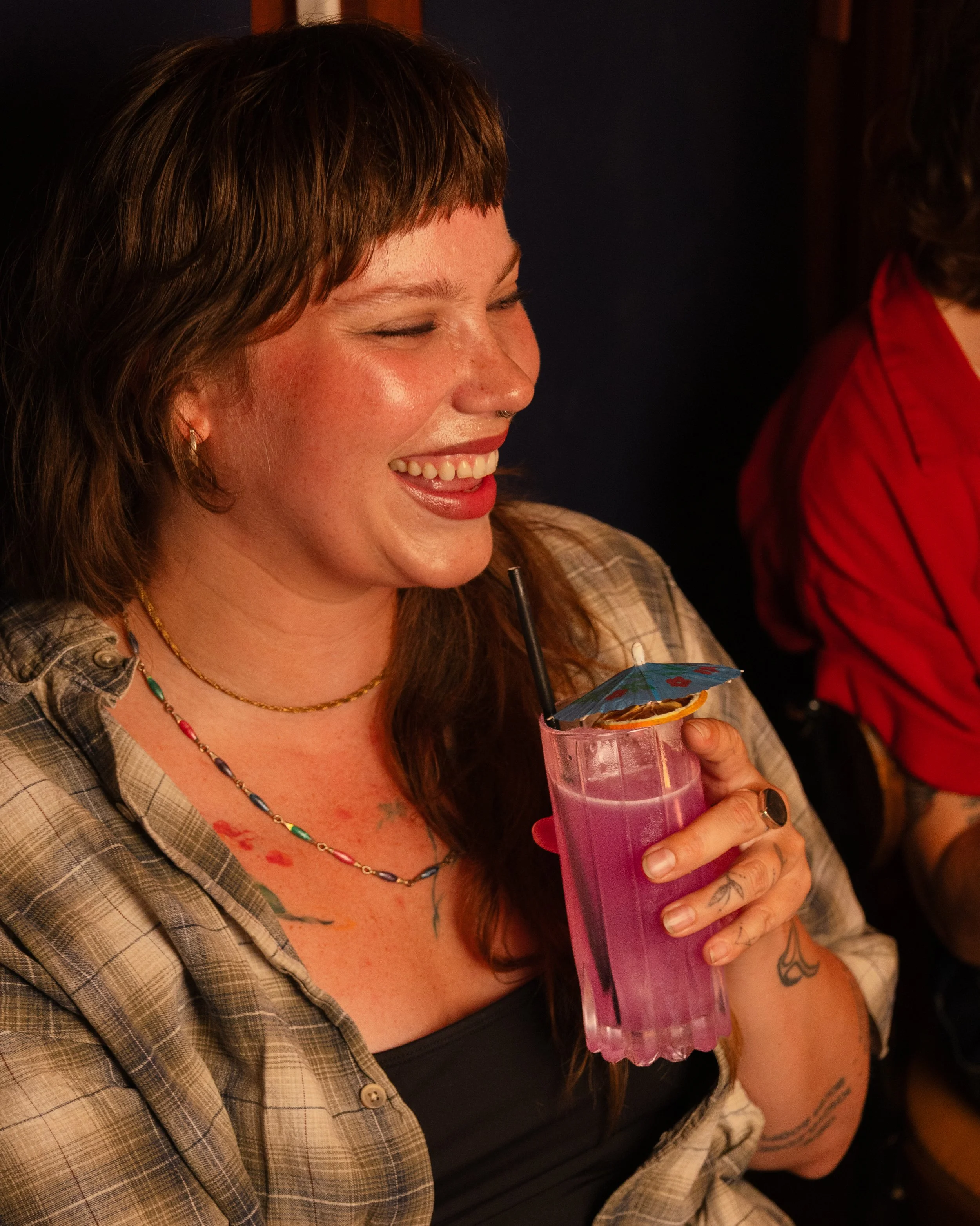 A woman with red hair, tattoos, and a colorful necklace holding a pink cocktail with a straw and orange slice, smiling happily.