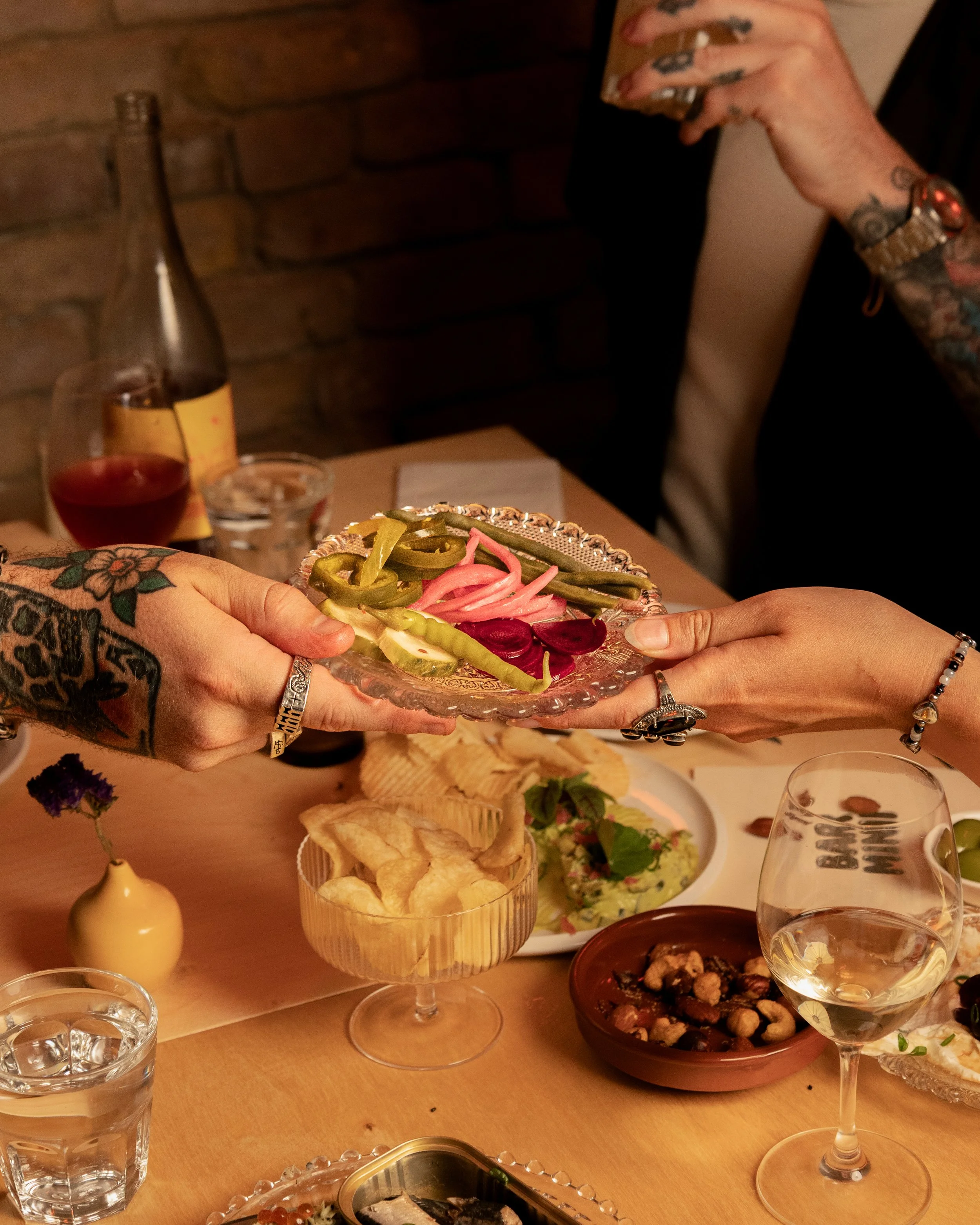 Two people exchanging a plate of pickled vegetables at a dinner table with various dishes, wine, and a bottle in a cozy, brick-walled restaurant.
