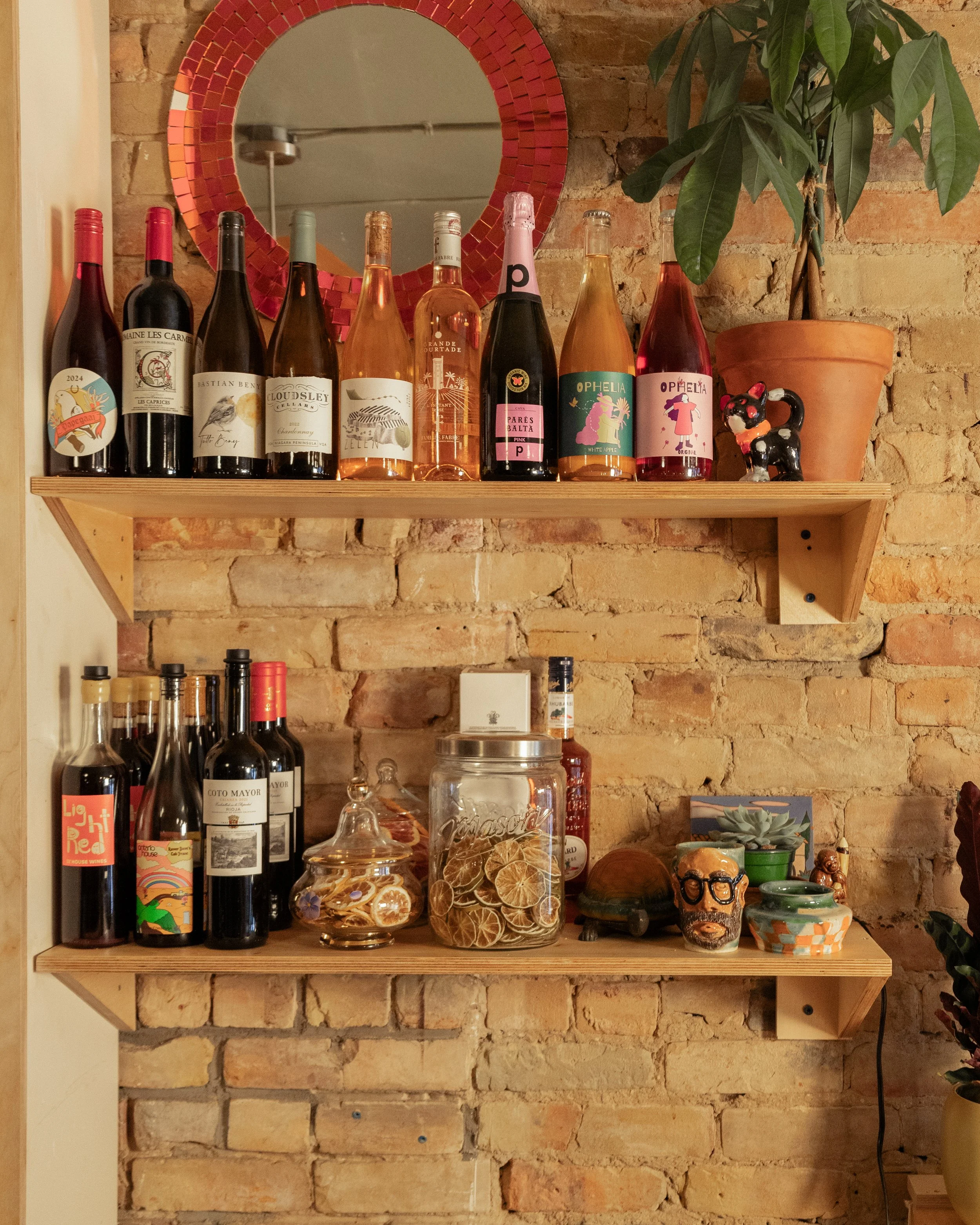Two wooden shelves mounted on a brick wall, holding wine bottles, decorative jars, a potted plant, and small decorative items including a black cat figurine, a mask, and ceramic pots.