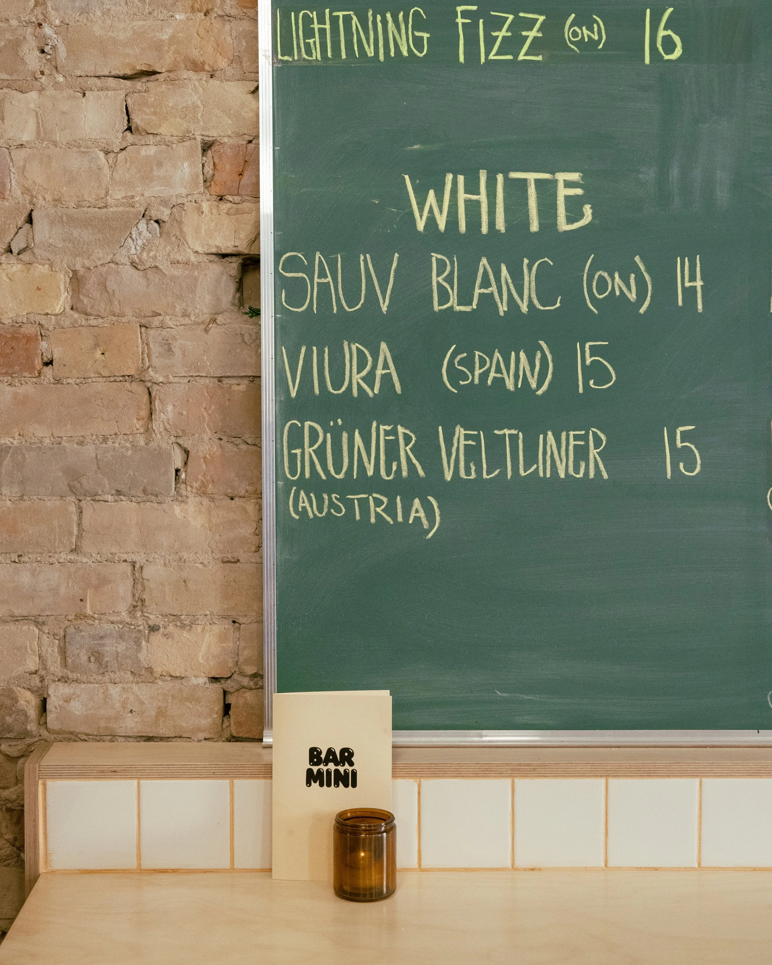A green chalkboard menu displaying types of white wine with prices, situated next to a brick wall, with a small amber glass jar and a cream-colored menu card labeled 'Bar Mini' on a light wood counter.