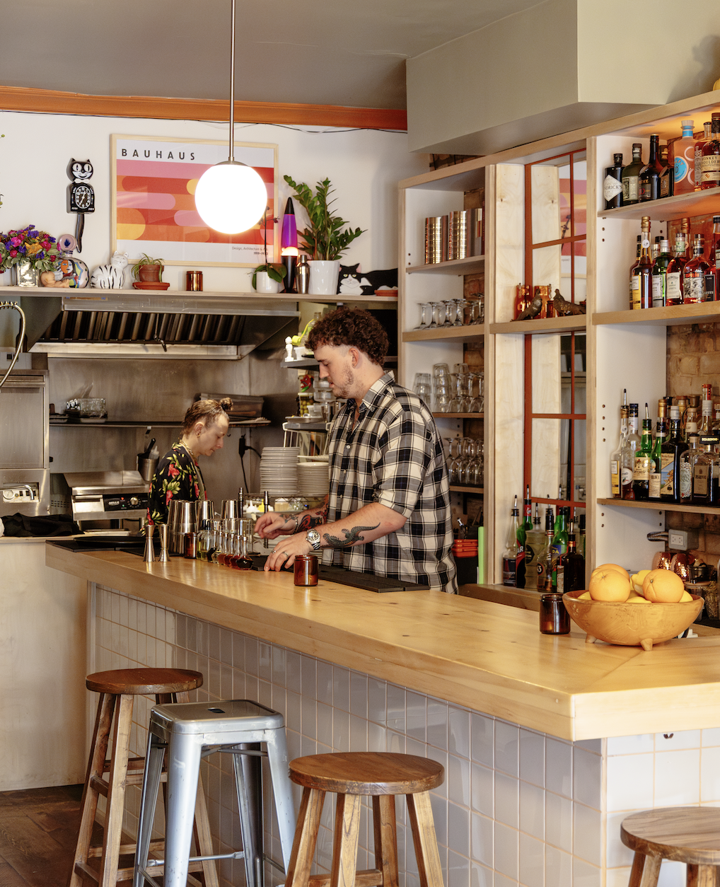 Inside a cozy bar or cafe with two bartenders preparing drinks, shelves stocked with liquor bottles, and modern decor including a framed Bauhaus poster and colorful plush toys.