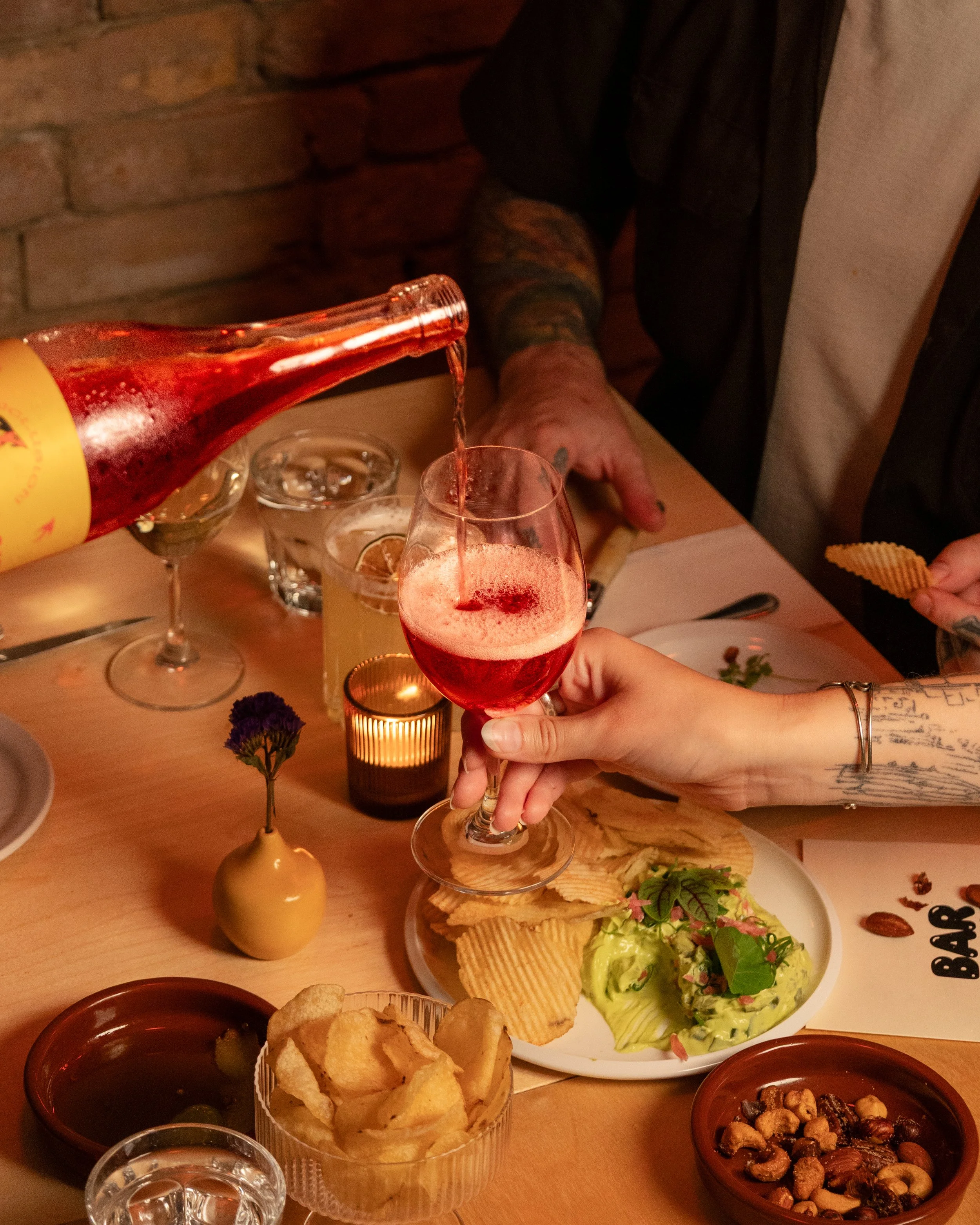 Person pouring a pinkish red drink into a wine glass at a table with snacks, a small vase, and candles, and a partially visible tattooed arm.