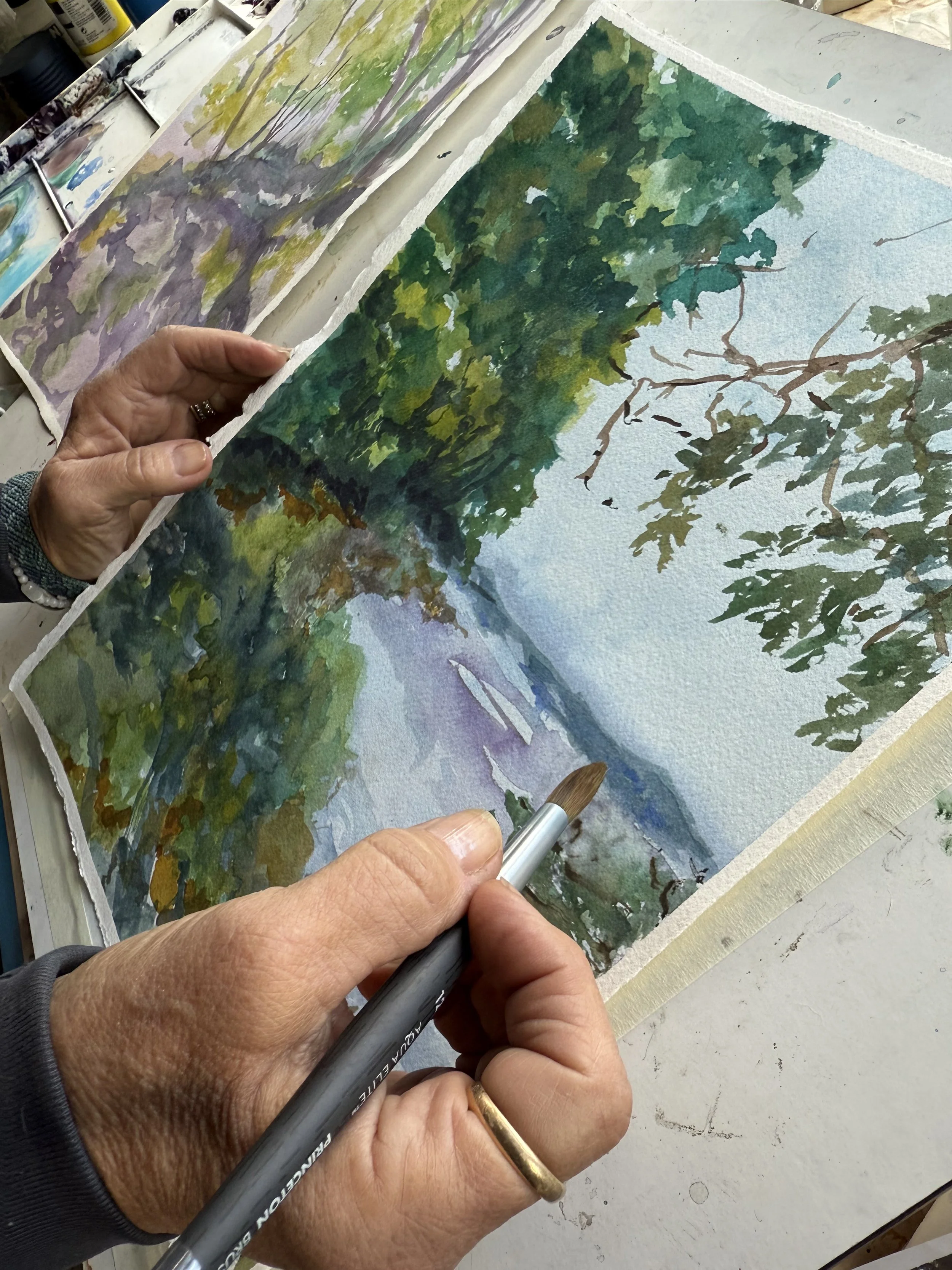 A person painting a watercolor landscape of a river flowing through a forest with leafy trees on the riverbank.