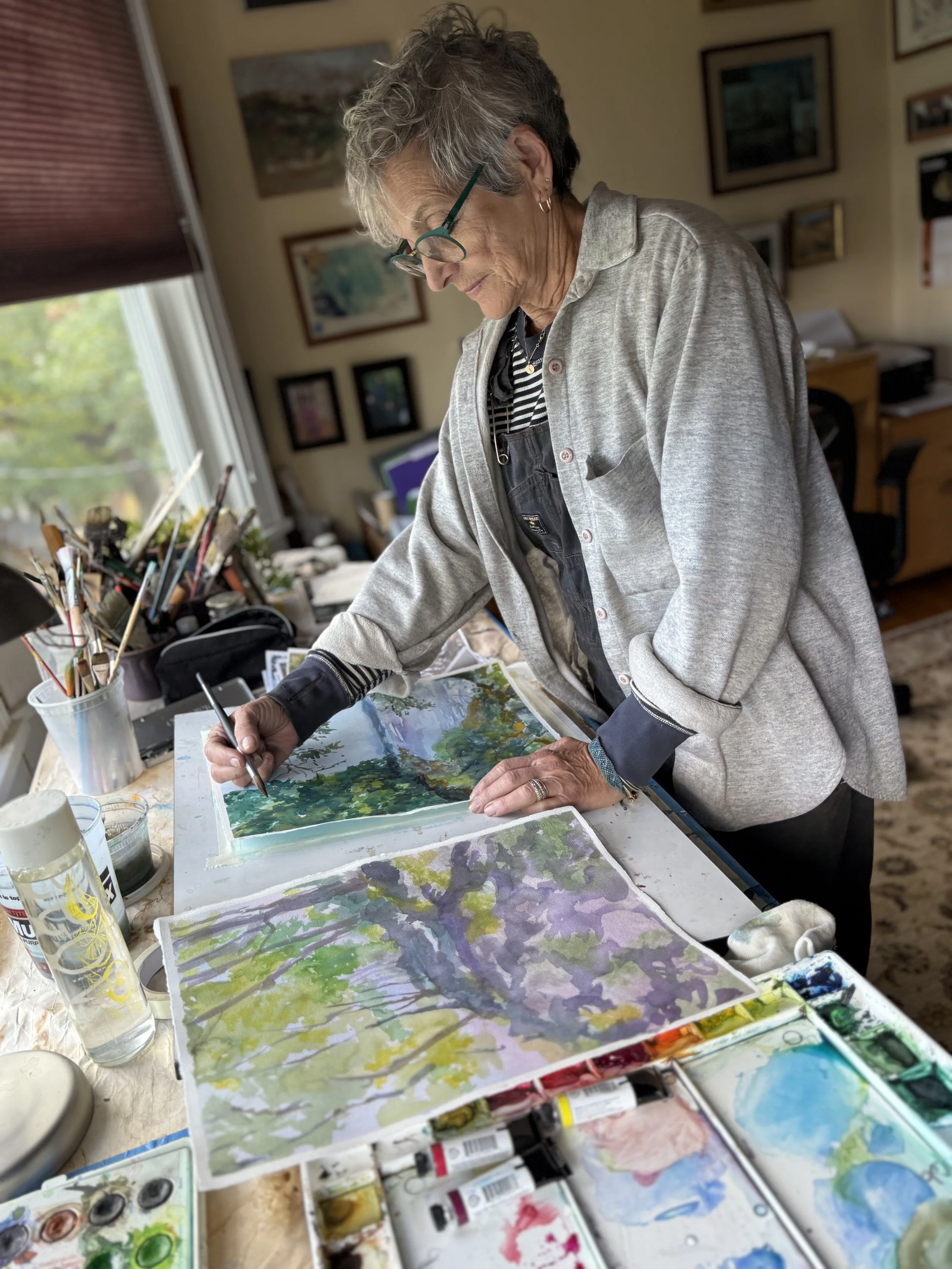 An elderly woman with short gray hair, glasses, and earrings is painting watercolor landscapes in an art studio. She is leaning over a table with various art supplies, including watercolors, brushes, and paper, with paintings of trees and nature scenes around her.