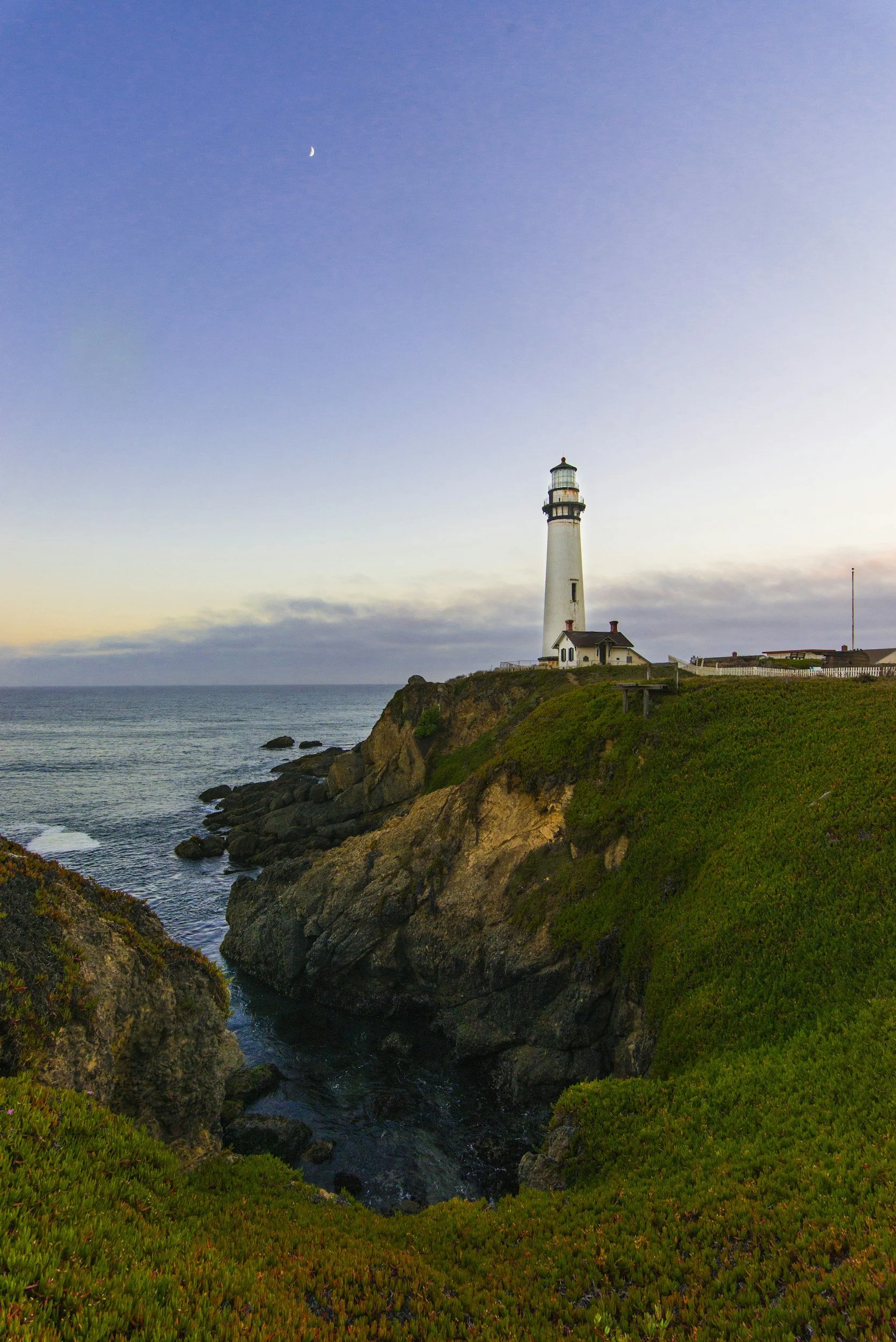 A lighthouse on a cliff overlooking the ocean at sunset with a clear sky and a crescent moon.