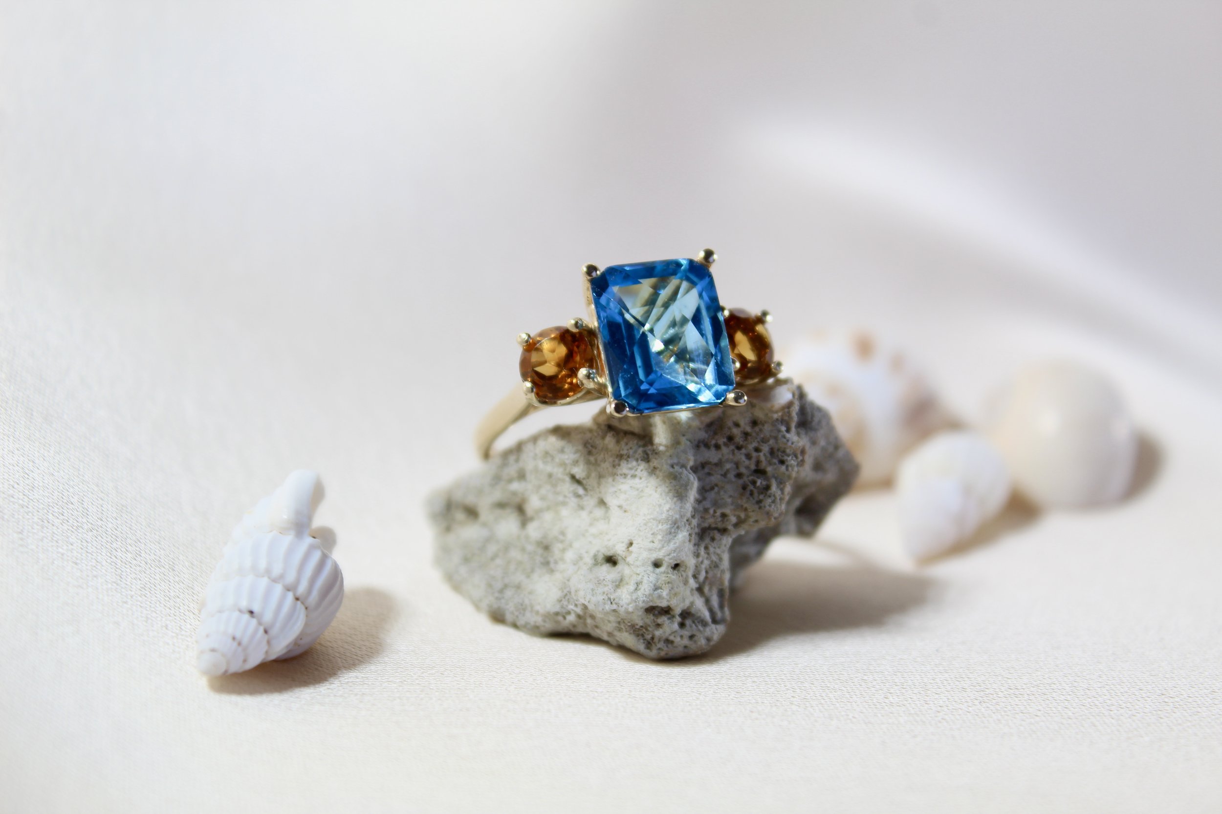 A ring with a large rectangular blue gemstone and two smaller orange gemstones, placed on a piece of coral with seashells around it.