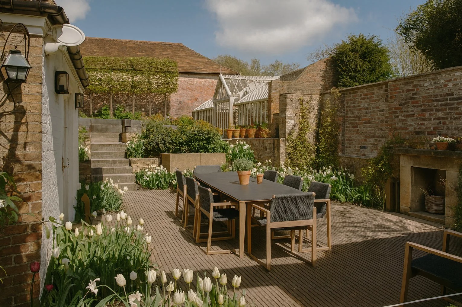 A charming courtyard garden with tulip-filled borders, a long outdoor dining table, and a brick wall backdrop with climbing plants and a greenhouse in the background.