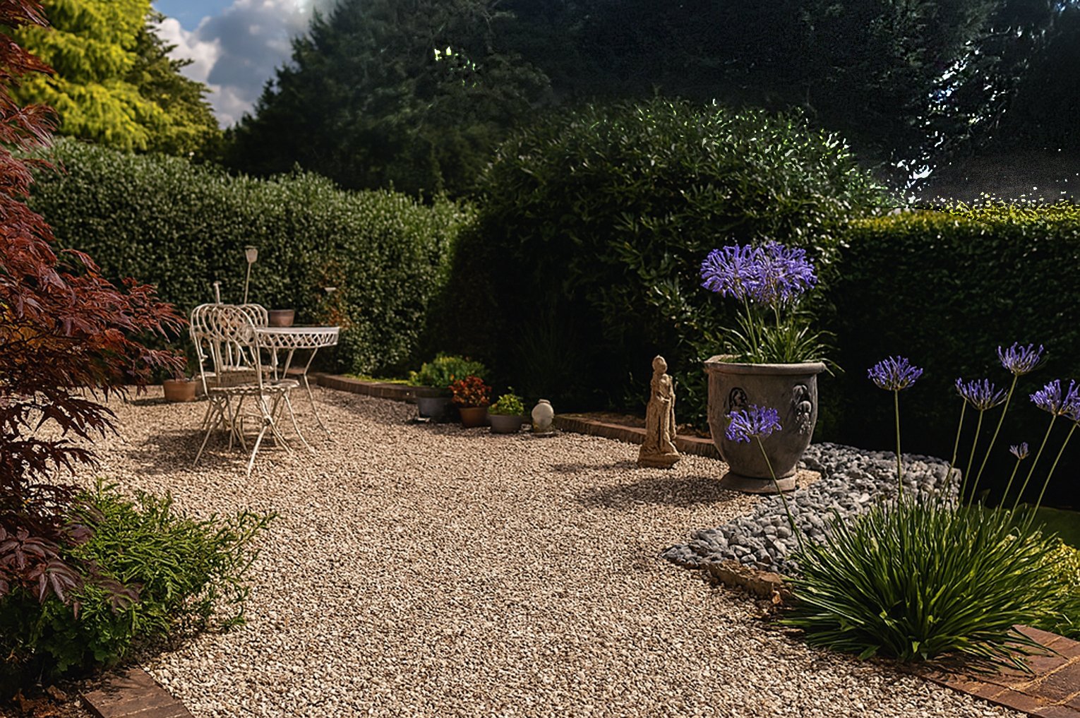 Sunny garden seating area with white chairs, gravel pathway, potted plants, and cloudy blue sky.