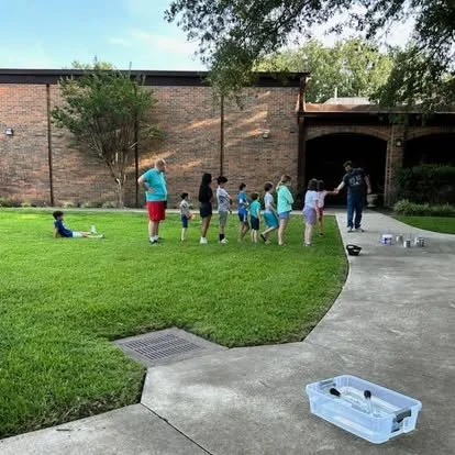Children and adults standing in line outdoors on a grassy area near a brick building, participating in a game or activity, with a small plastic container and bottles on the ground nearby.