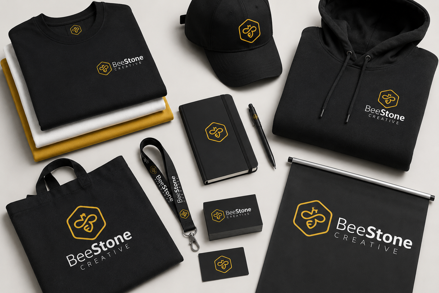 Collection of branded merchandise items including black T-shirt, black baseball cap, black hoodie, black tote bag, black notebook, business card, lanyard, flag, and sticker, all featuring the BeeStone Creative logo in yellow and white.
