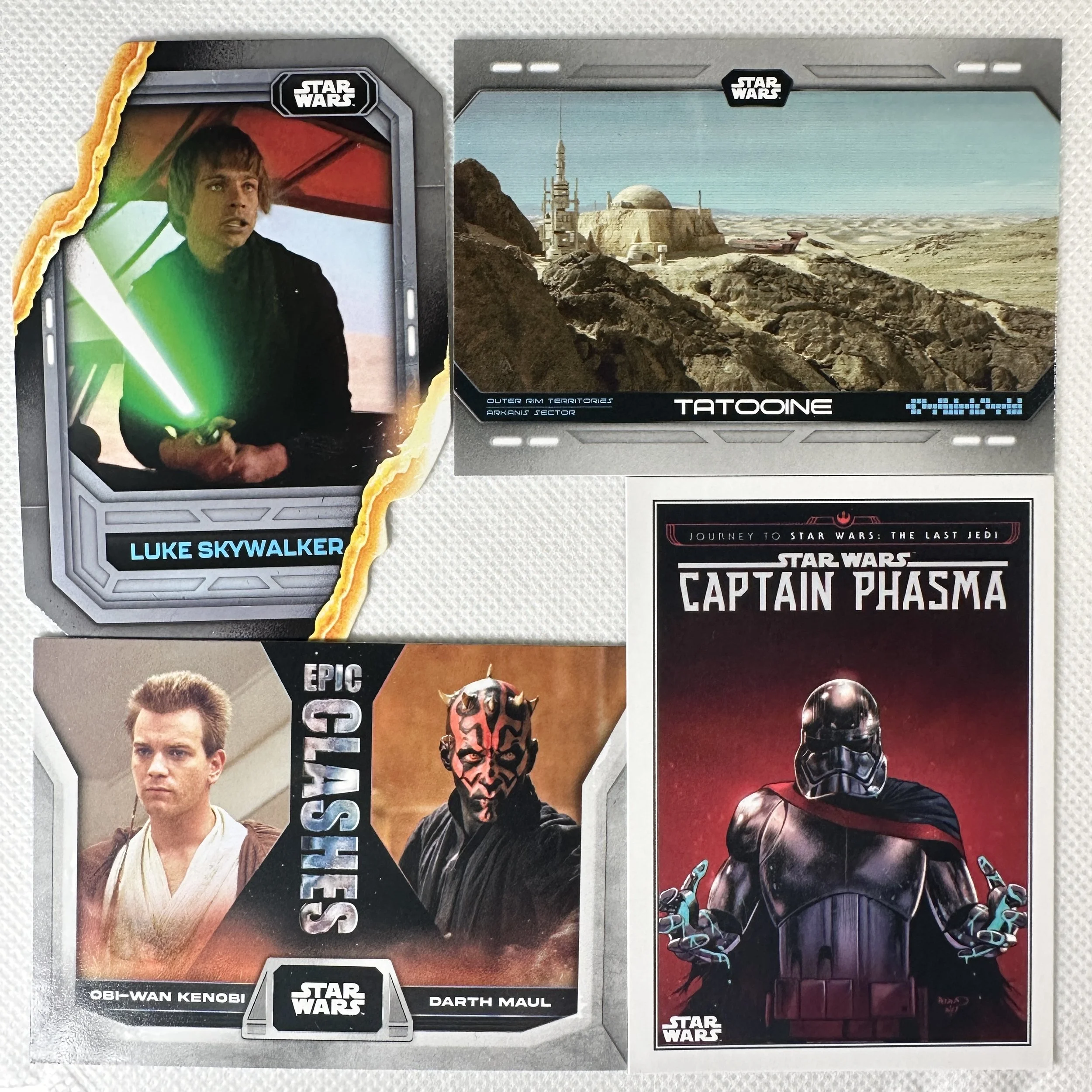Four Star Wars trading cards laid out on a surface. The top left card features Luke Skywalker holding a green lightsaber, with a torn edge design. The top right card depicts the Tatooine landscape with a city, mountains, and a blue sky. The bottom left card shows Obi-Wan Kenobi and Darth Maul with the text 'Epic Showdown.' The bottom right card features Captain Phasma in a black suit and helmet, with arms outstretched. All cards have Star Wars branding.