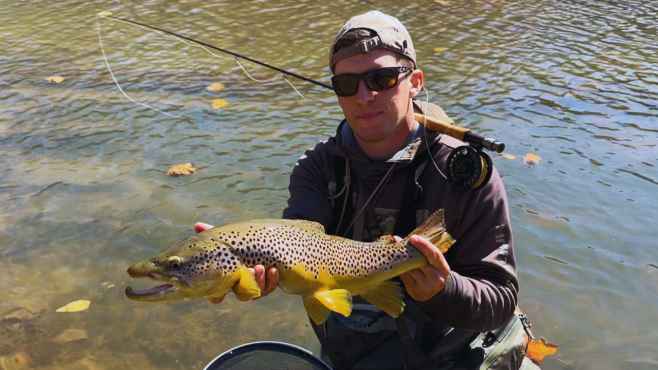 Man wearing sunglasses and a cap, holding a large spotted fish by a river with fallen leaves, with fishing gear visible.