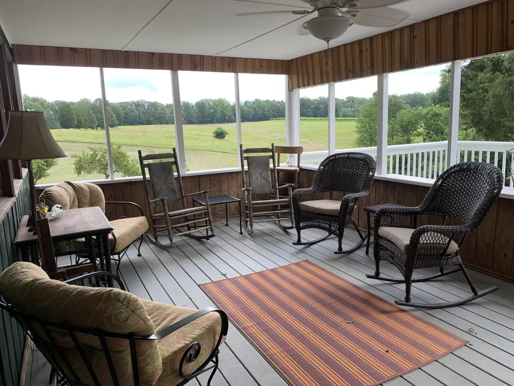 Covered porch with wooden walls and ceiling fan, furnished with rocking chairs, wicker chairs, a sofa, a side table, a lamp, outdoor rug, overlooking a lush green field with trees in the background.