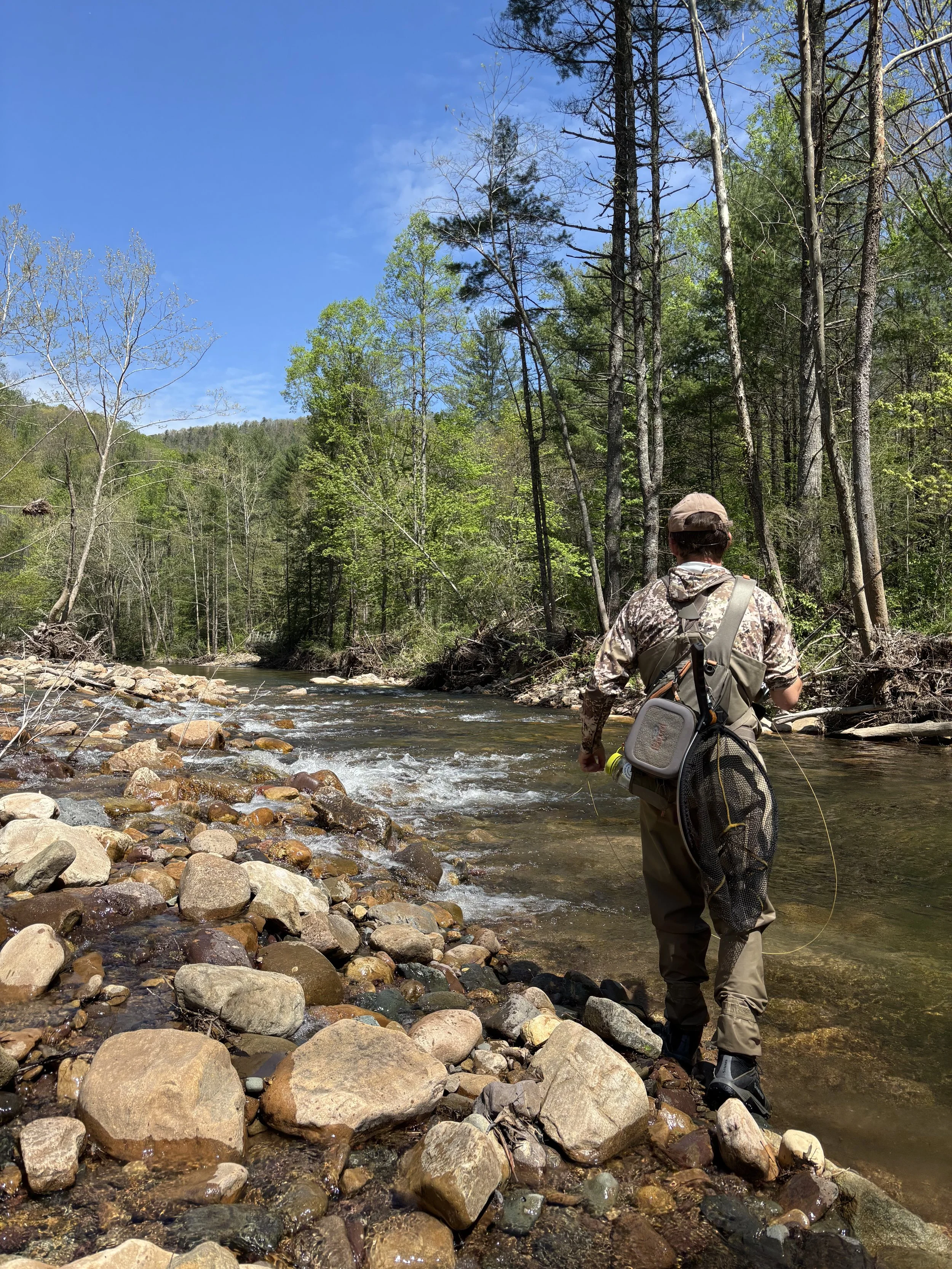 Person wearing outdoor gear and fishing at a rocky stream in a forest with a clear blue sky.