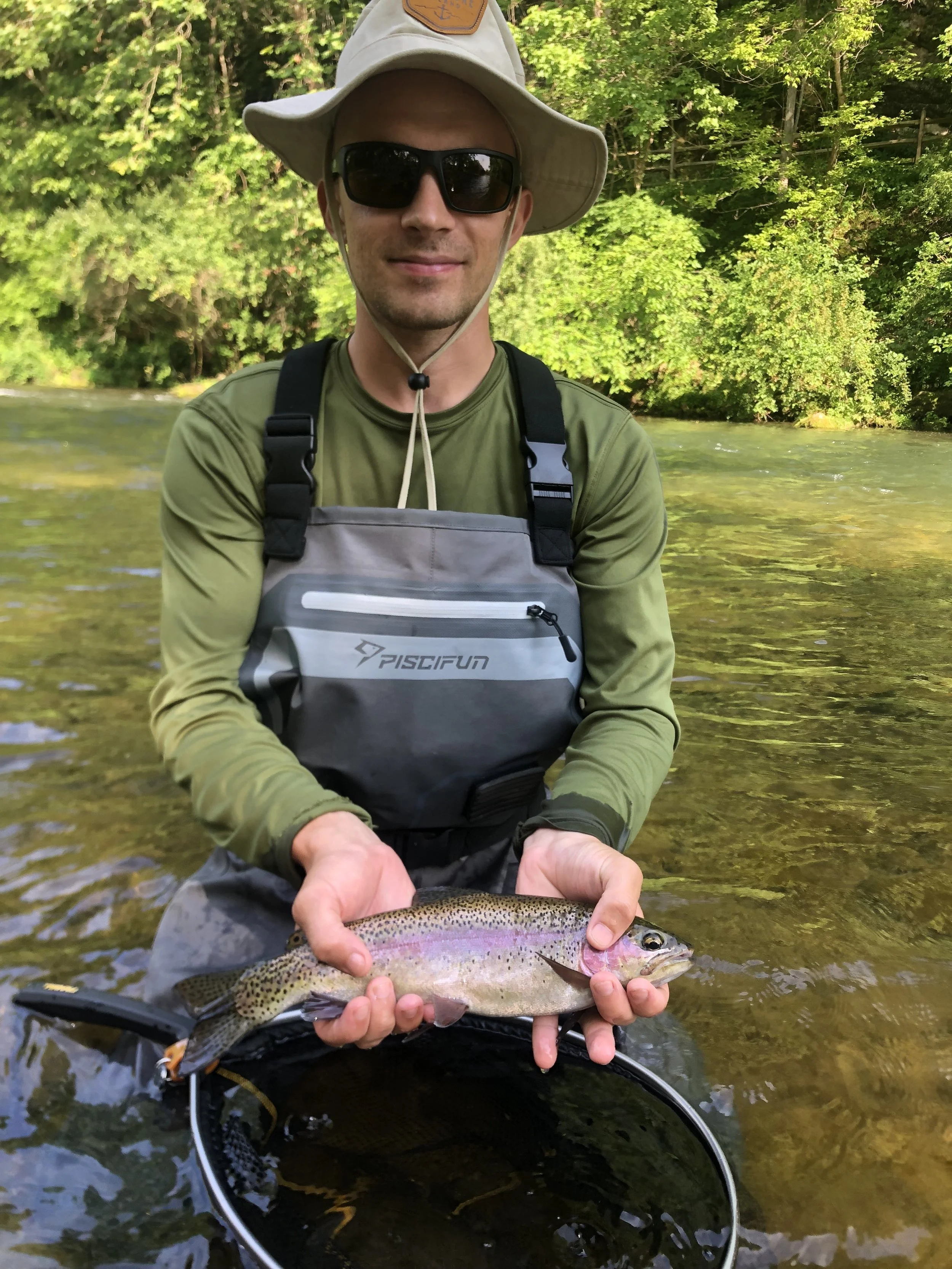 A man wearing sunglasses, a wide-brimmed hat, and a green shirt stands in a stream, holding a large rainbow trout with spots. He is smiling and appears to be proud of his catch, with dense green foliage in the background.