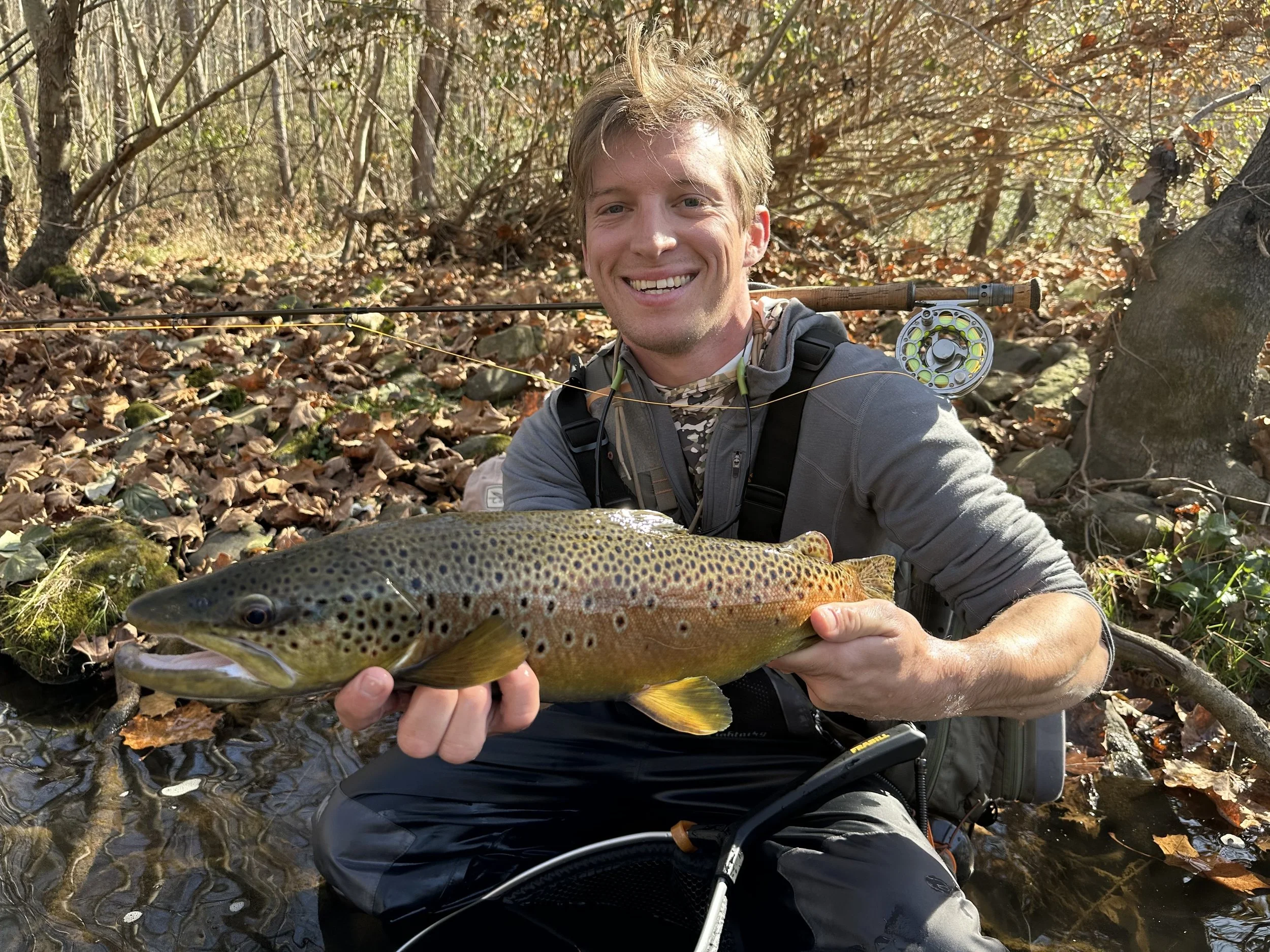 A man smiling while holding a freshly caught rainbow trout in a forest setting with fallen autumn leaves and a fishing rod across his shoulder.