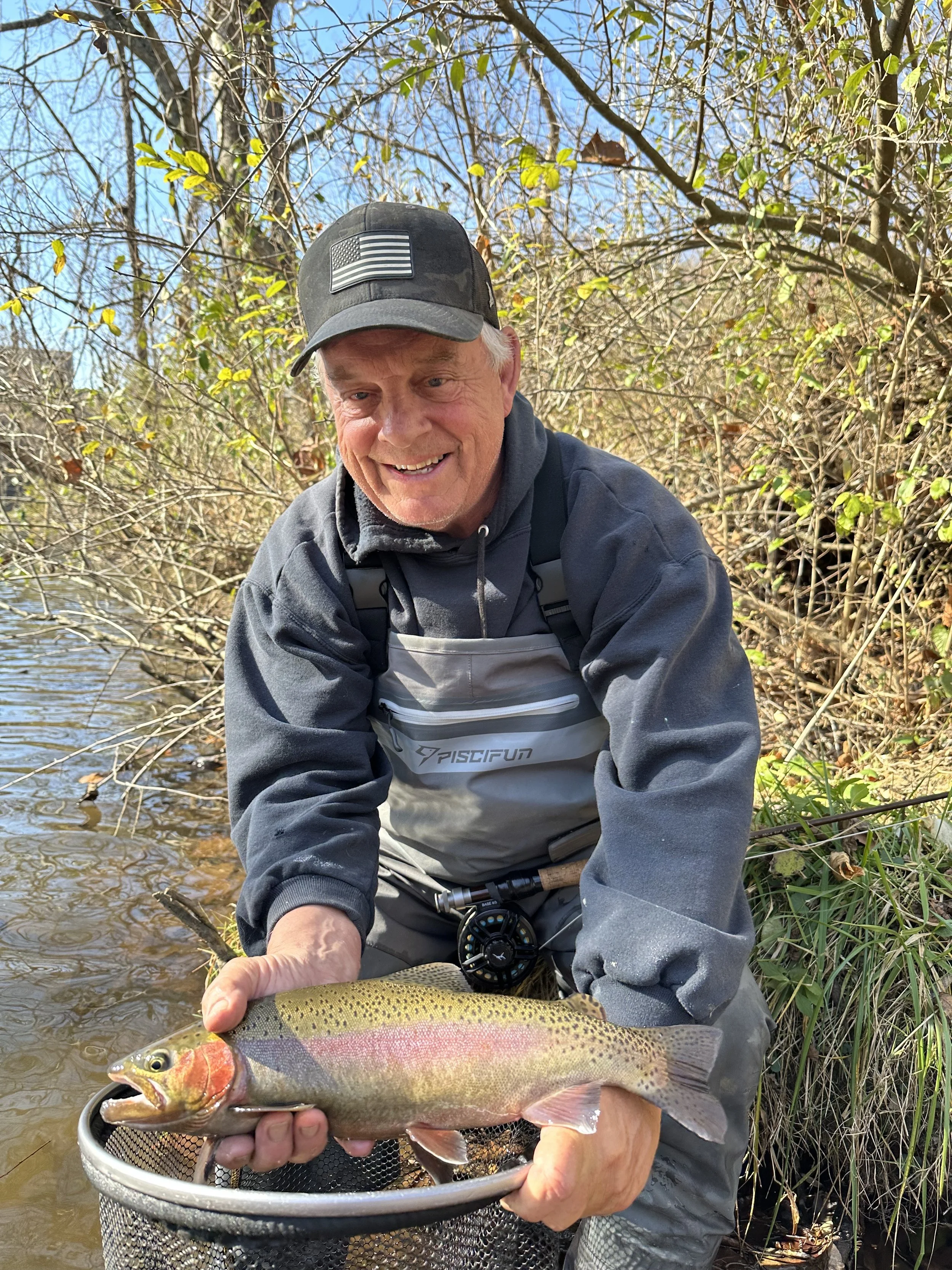 A man wearing a gray jacket and a black cap with a U.S. flag patch is smiling and holding a large rainbow trout over a fishing net in a river, surrounded by leafless trees and bushes on a sunny day.