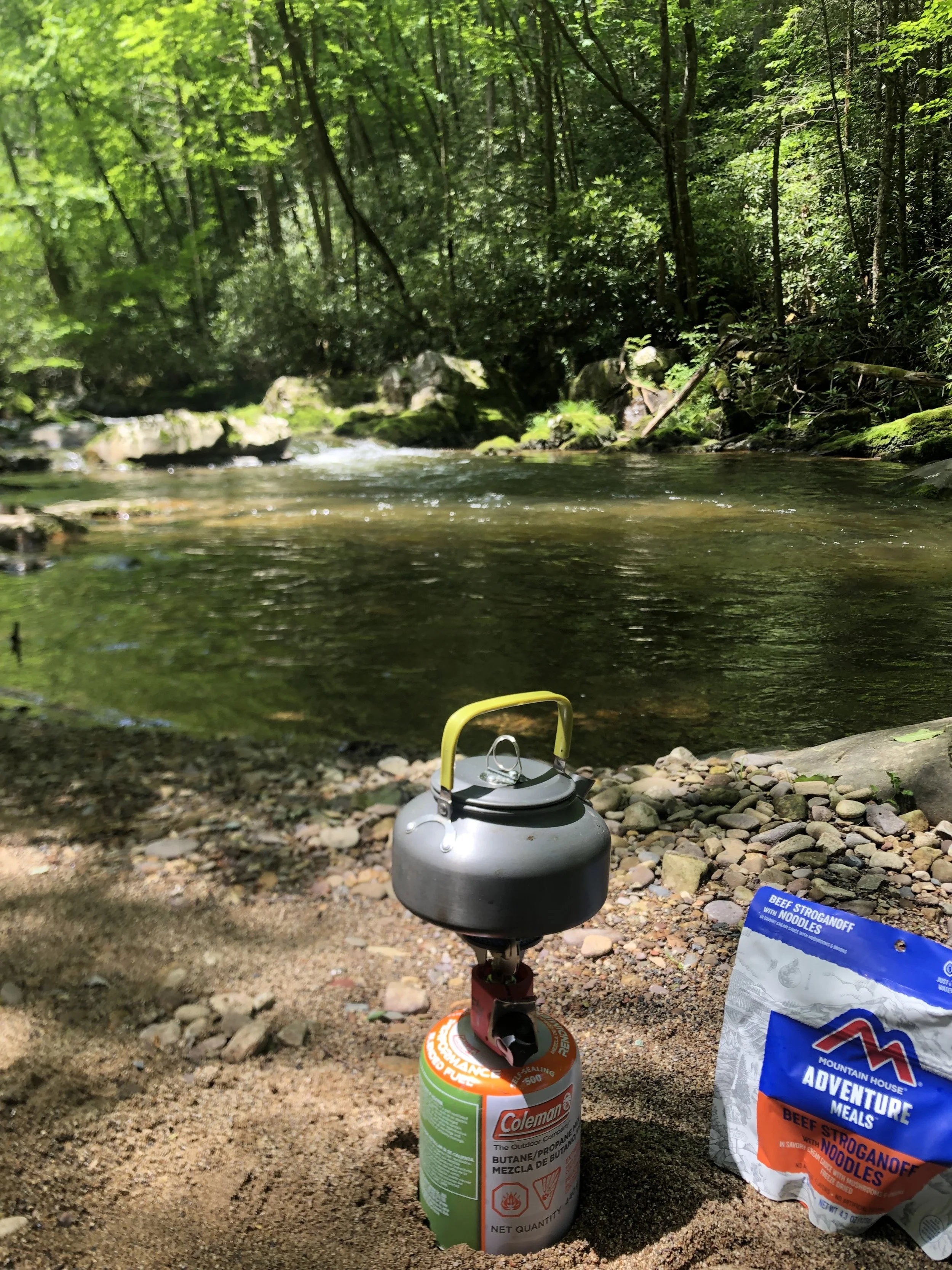 A camping stove with a gas canister and a kettle on top, set on a rocky ground near a stream in a forest. A bag of Mountain House adventure meals and a package of beef stroganoff with noodles are nearby.