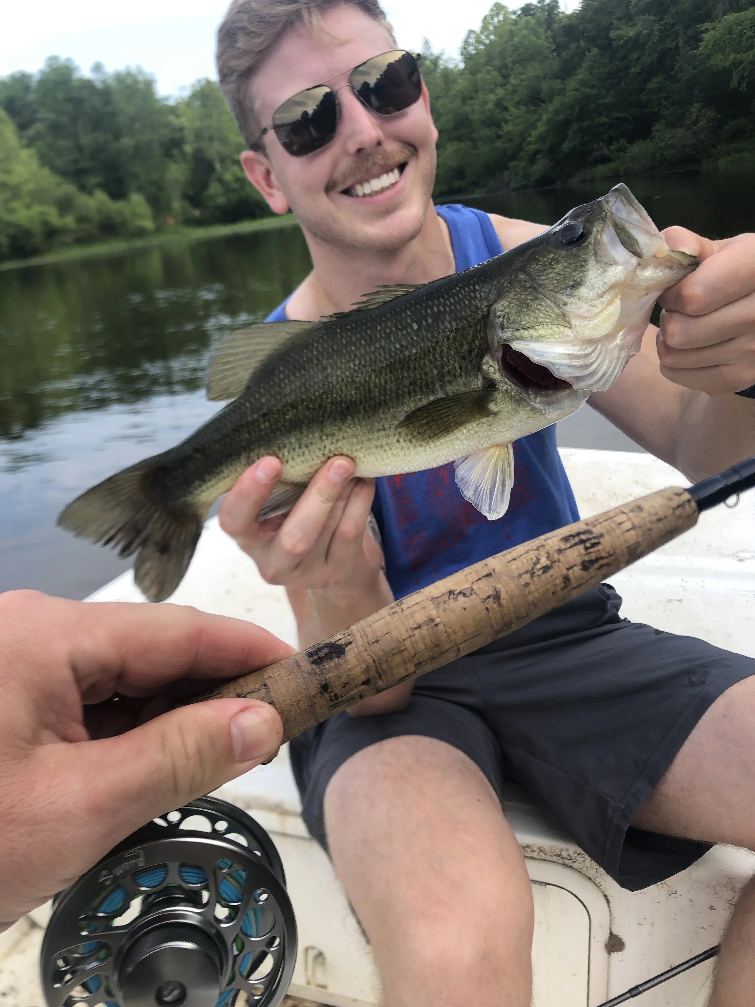 A man is smiling and wearing sunglasses while holding a largemouth bass fish on a boat during daytime with a background of water and green trees.
