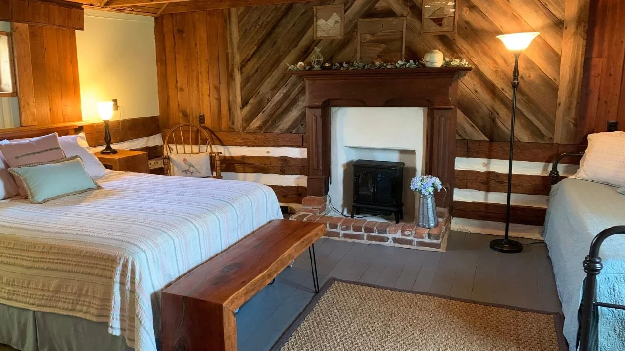 Cozy rustic bedroom with a wooden wall and brick fireplace, two beds with white and pastel bedding, a wooden bench, lamps, and decorative items on the mantle.