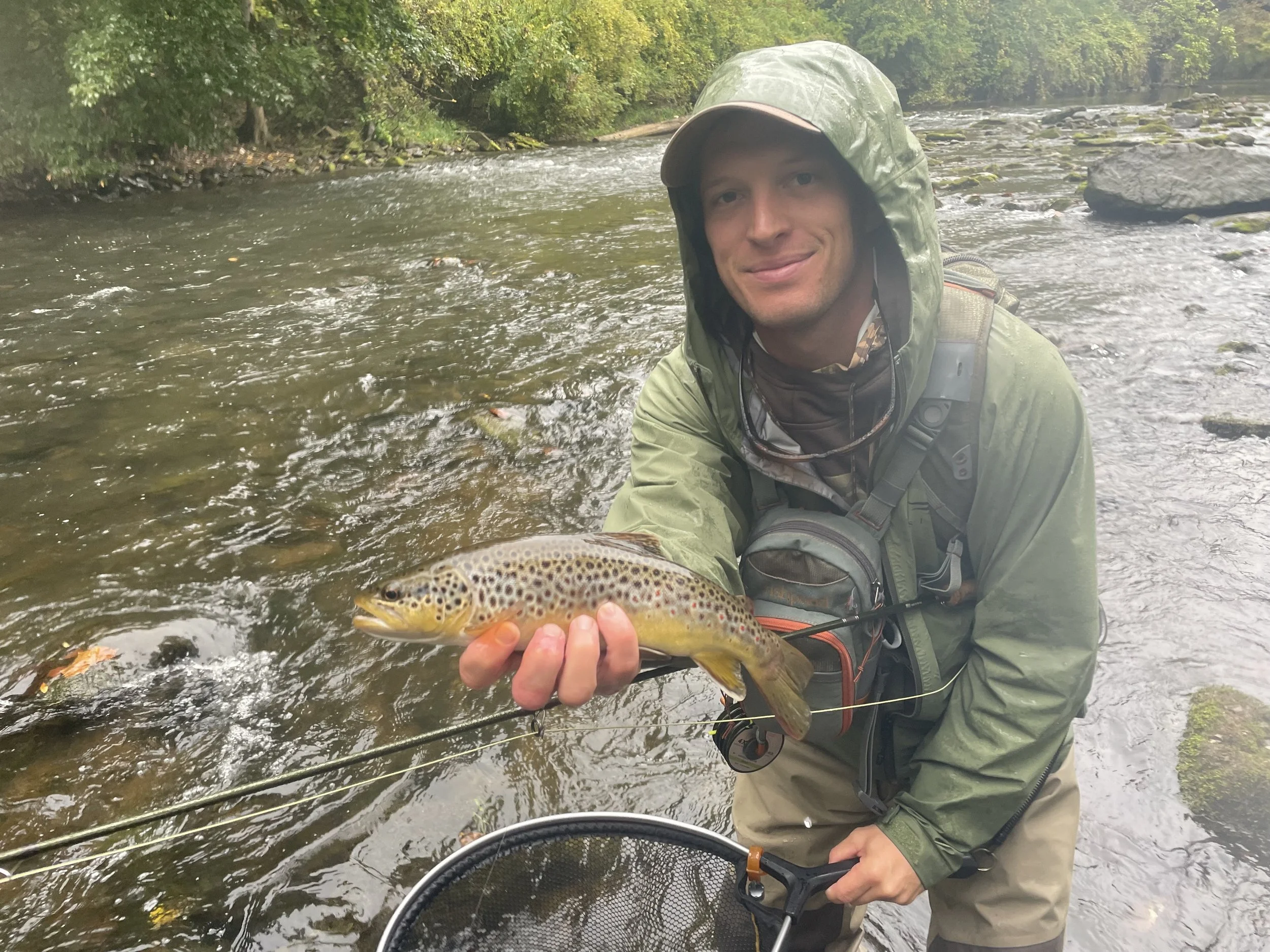 A man wearing outdoor gear and a hooded waterproof jacket, standing in a river, holding a freshly caught brown trout fish with a smiling expression.