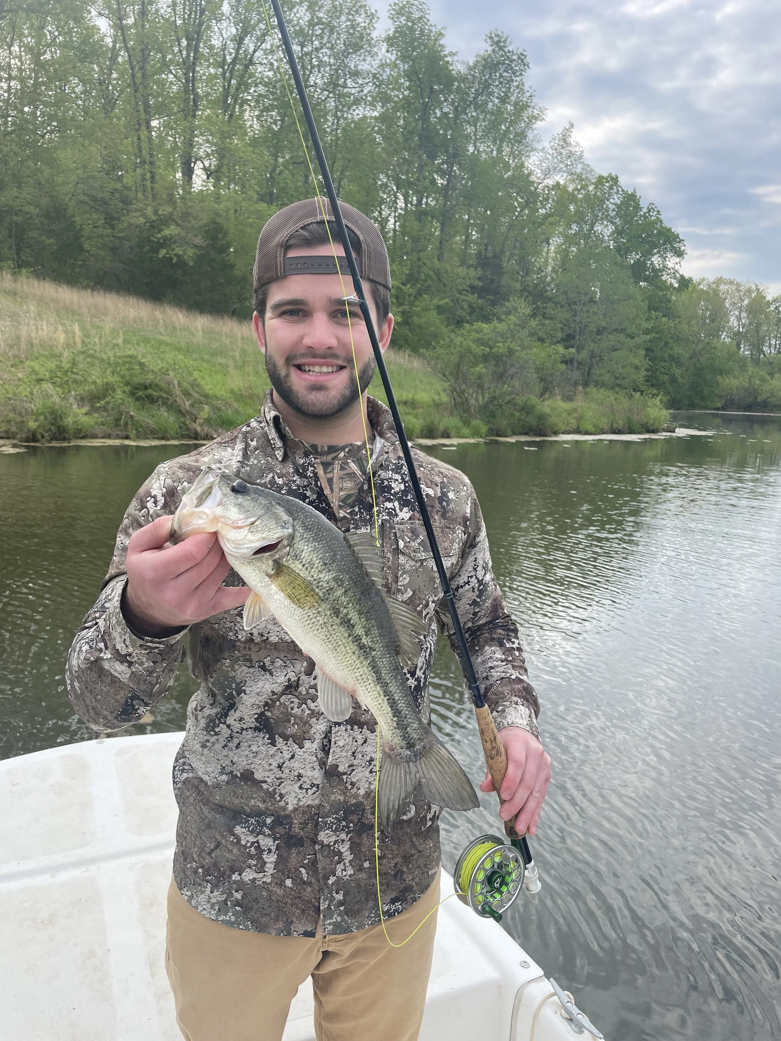 A man in camouflage shirt and tan pants on a boat holding a fish in one hand, with fishing gear in the other, near a body of water with trees and a cloudy sky in the background.
