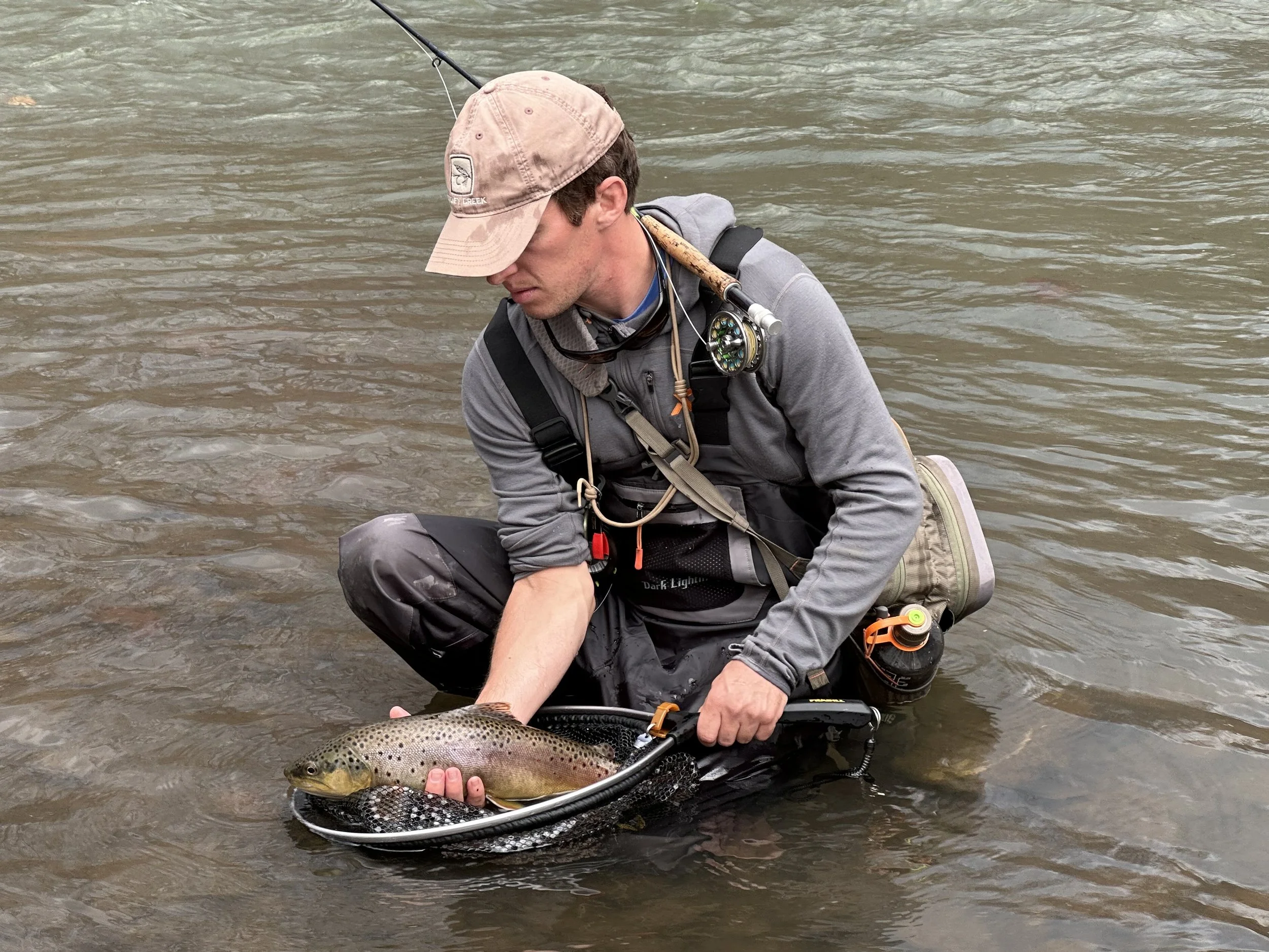 A man wearing outdoor fishing gear kneeling in a river, holding a large rainbow trout with both hands.