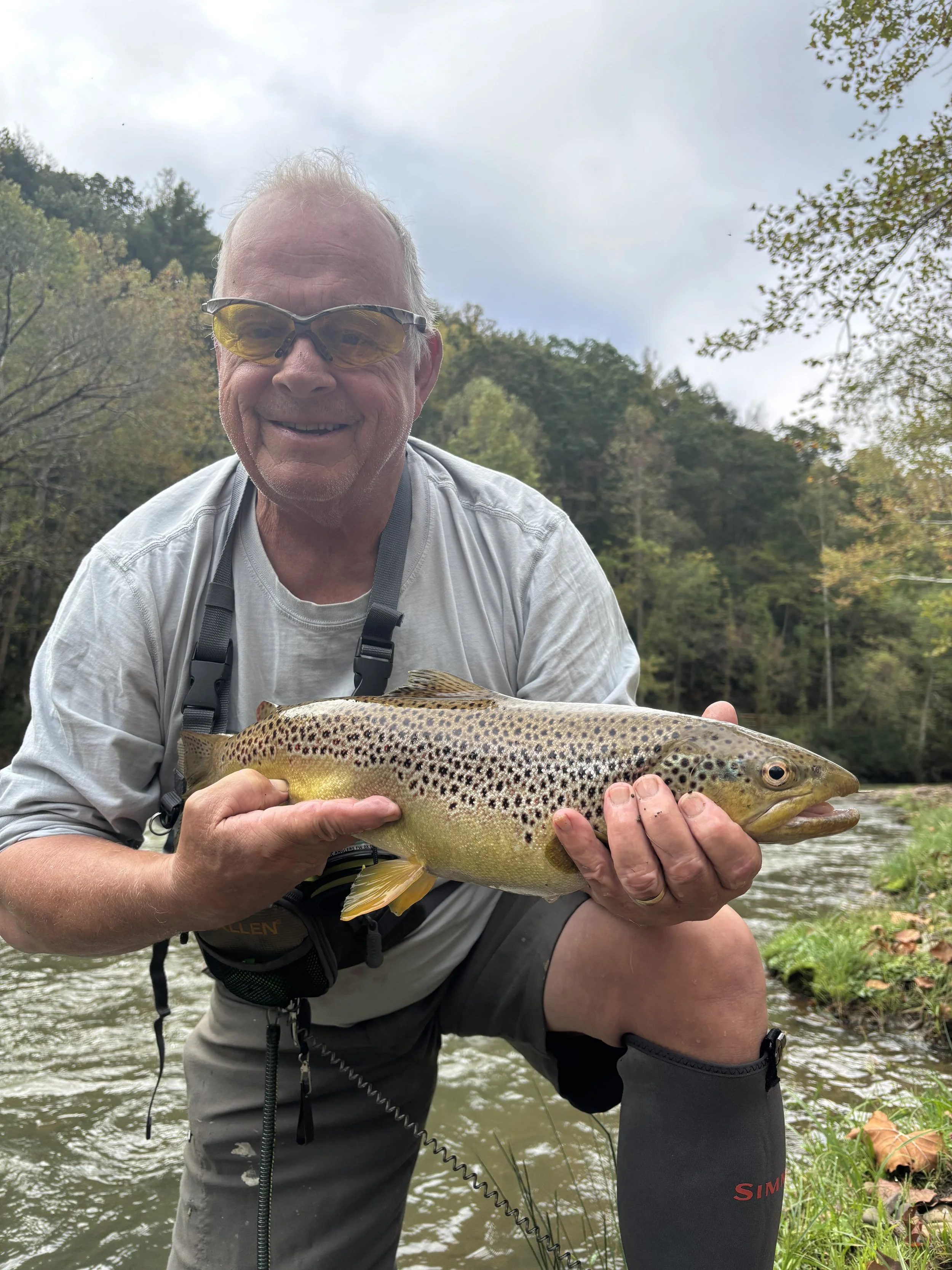A man holding a rainbow trout fish outdoors near a river with trees and cloudy sky in the background.
