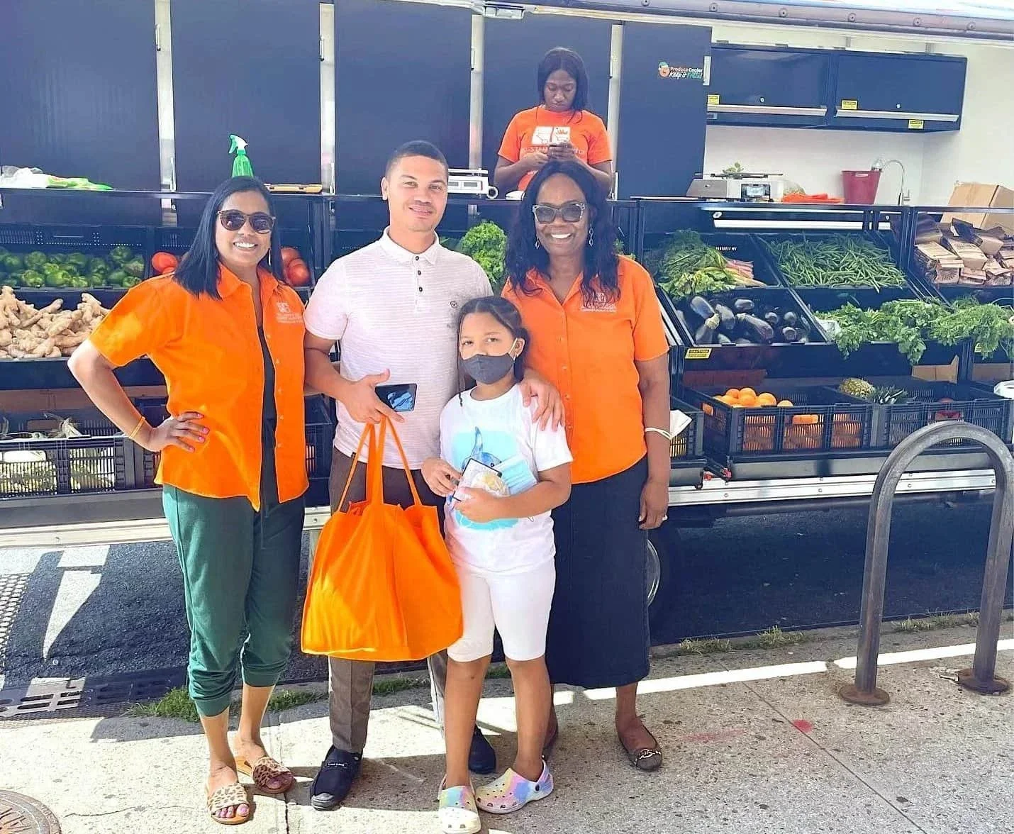 Group of five people standing in front of a produce truck at a farmer's market, with fresh vegetables like ginger, green peppers, broccoli, and cucumbers visible on the truck. Two women are wearing orange shirts, one woman is wearing sunglasses, one 
