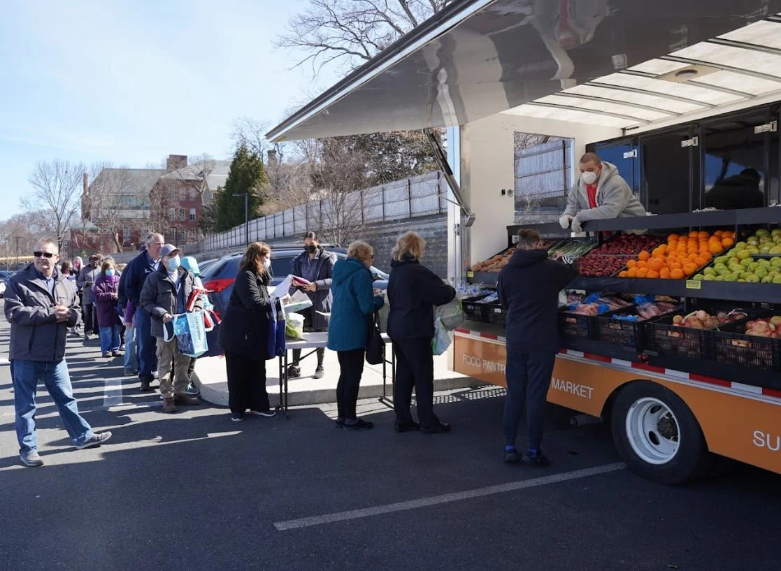 People standing in line at a mobile fresh produce market truck, buying fruits and vegetables, with some wearing masks, on a clear day.